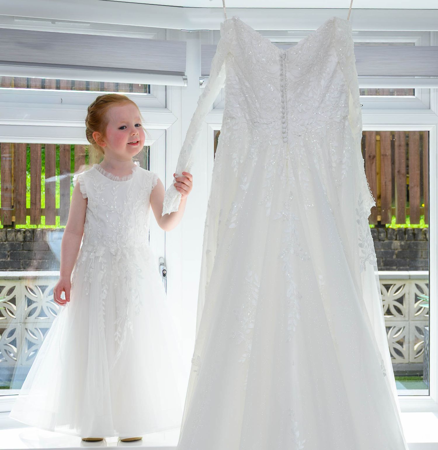 Young girl in white dress standing next to an elegant lace wedding gown hanging near a bright window.