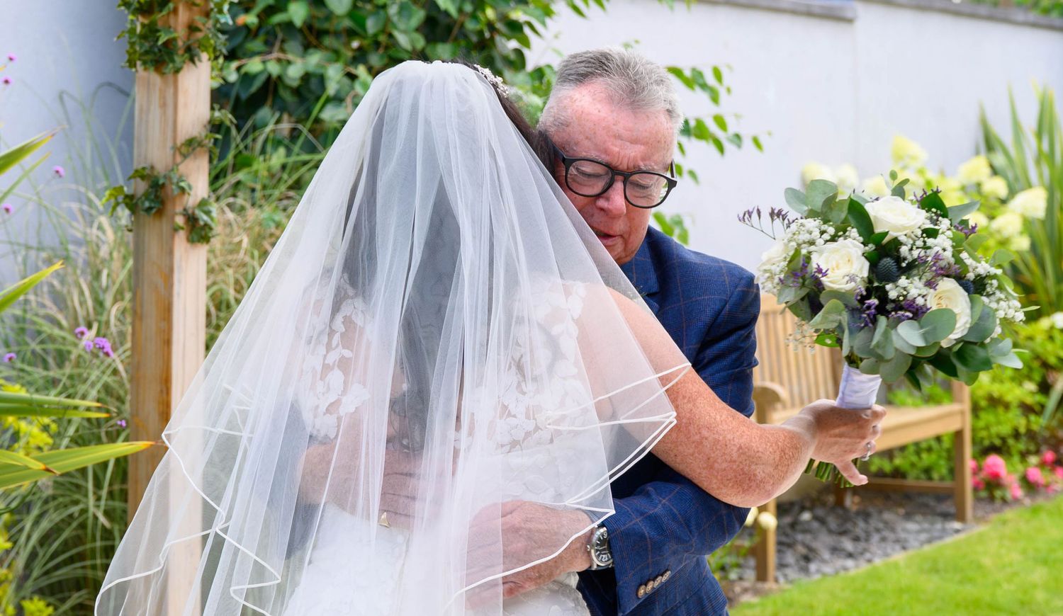 Bride in white gown and veil embracing older man in blue suit outdoors, holding white floral bouquet at wedding ceremony.