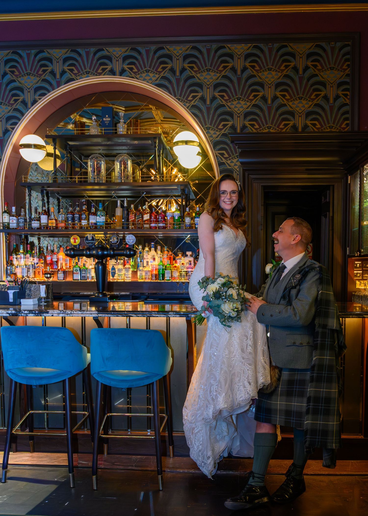 Bride and groom in kilt pose at ornate bar with blue stools and art deco decor at Scottish wedding venue.