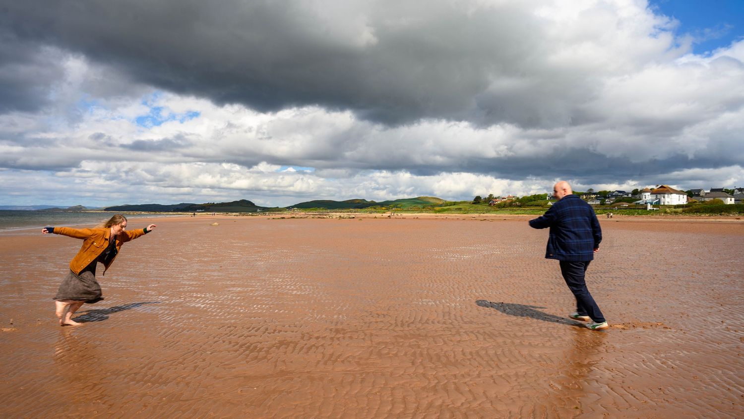 Two people playing on a vast red sand beach under dramatic cloudy skies near a coastal village.