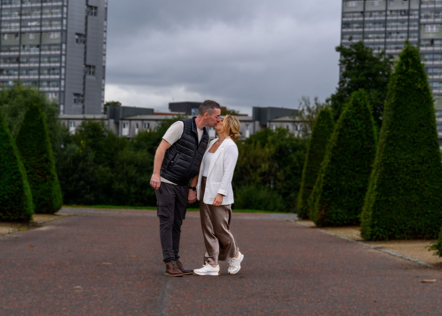 A couple walks together on a paved pathway through a garden with tall buildings visible in the background. Glasgow Green