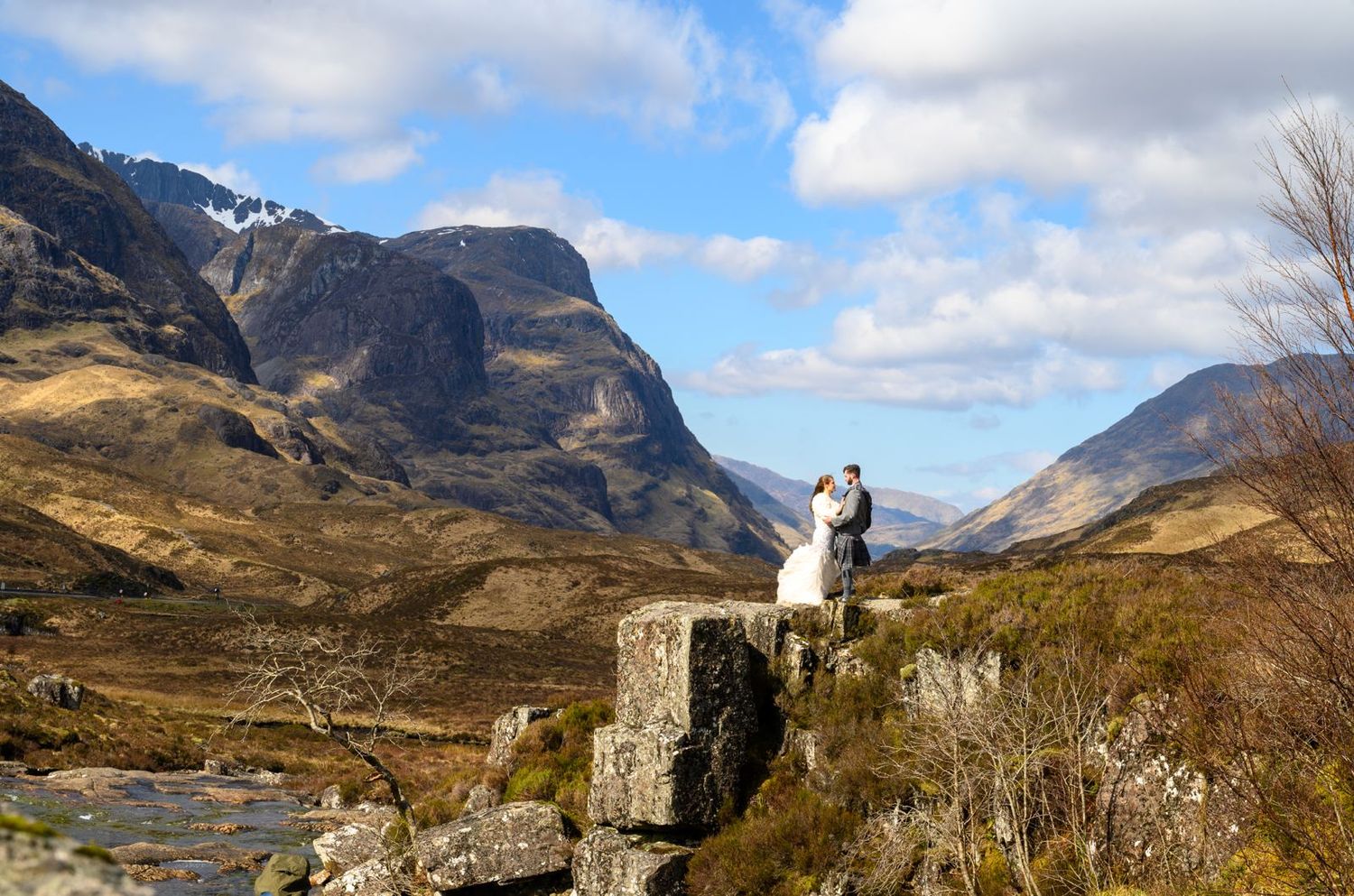 Wedding couple stands on ancient ruins overlooking a breathtaking Scottish Highland mountain valley.