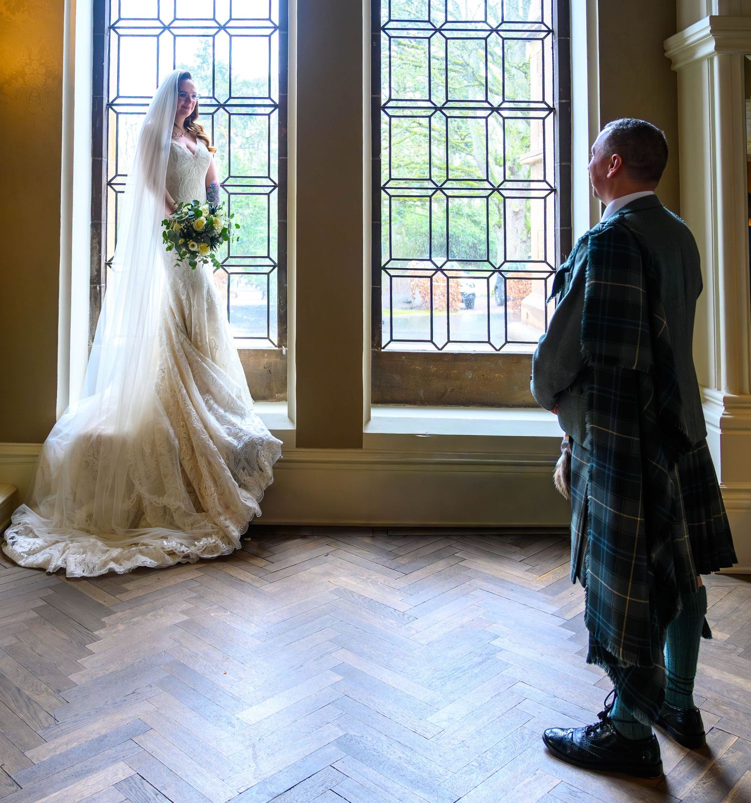 Bride in white gown and veil stands by tall window as groom in tartan kilt admires her in elegant venue.