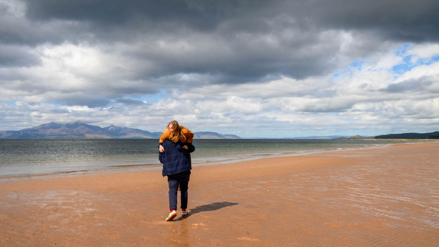A lone person with red hair walks along a sandy beach under dramatic cloudy skies with mountains in the distance.