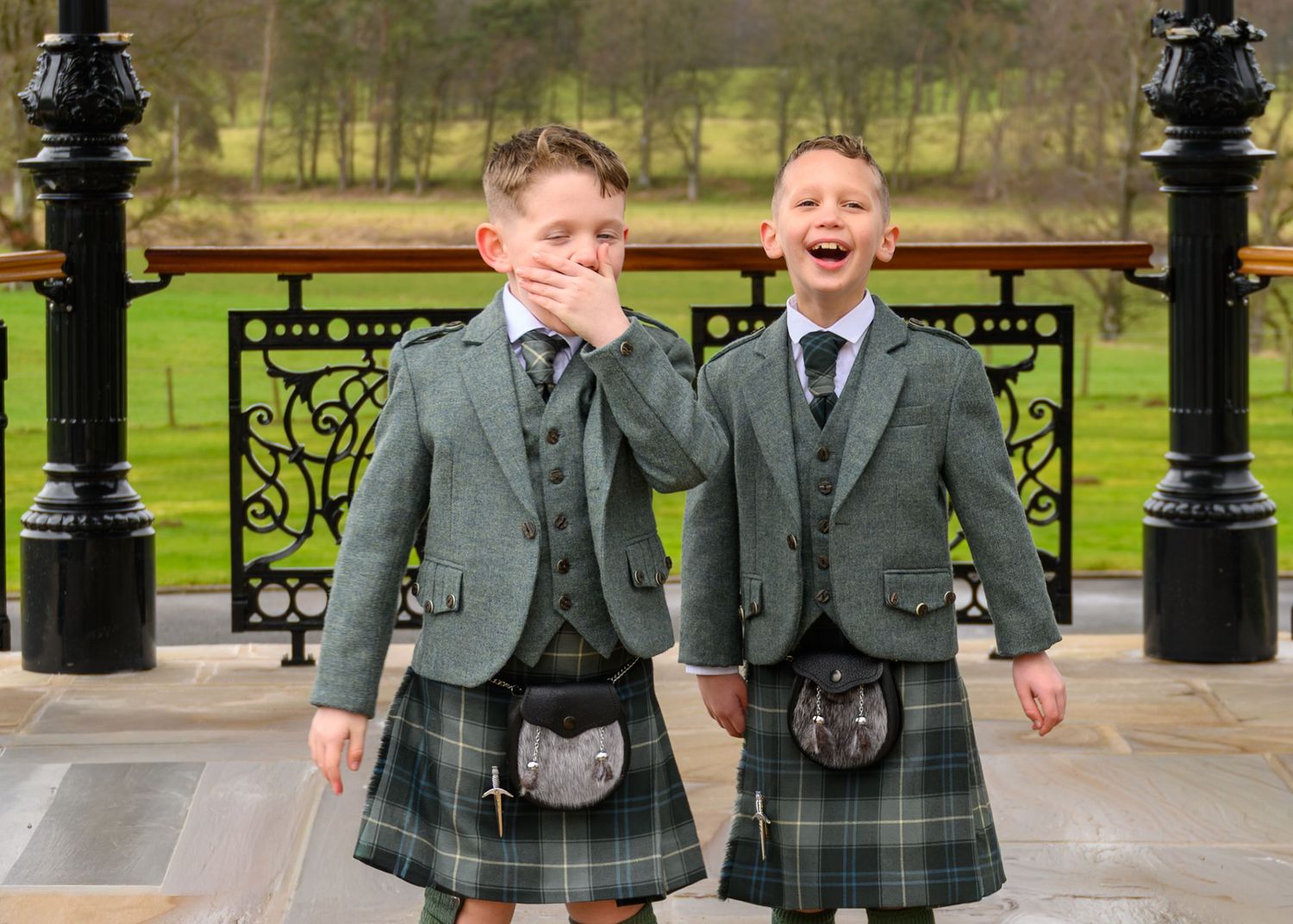 Two boys wearing traditional Scottish kilts and jackets laugh together outdoors at a formal venue.