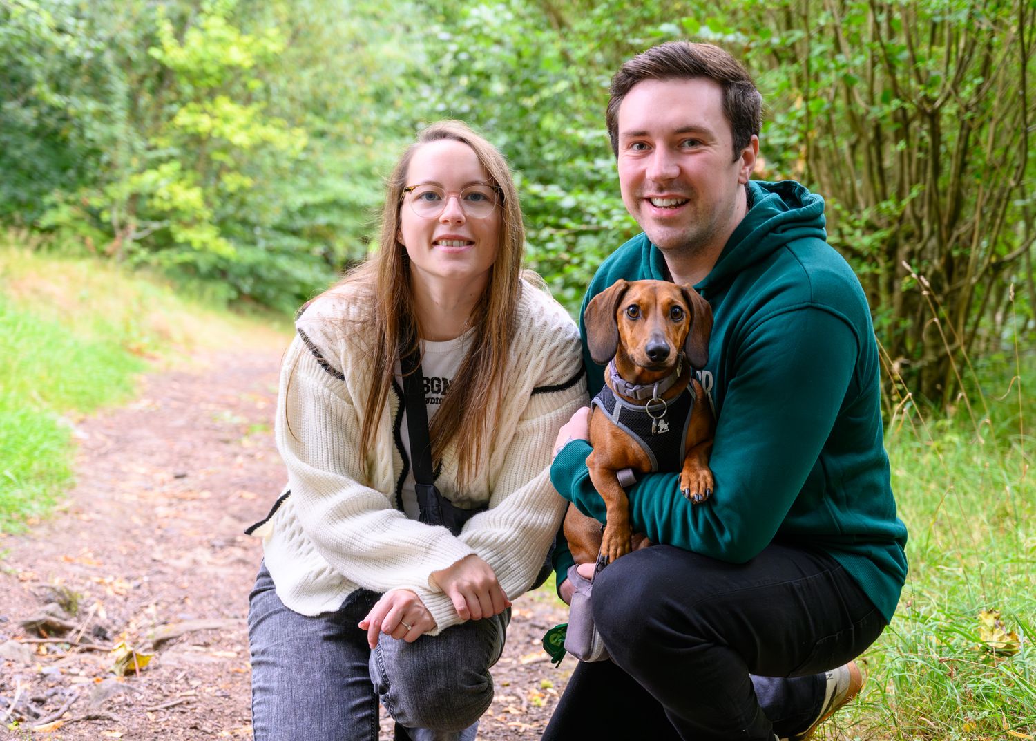 A couple in casual sweaters poses on a trail with their small brown dachshund puppy Freddy at Queens Park
