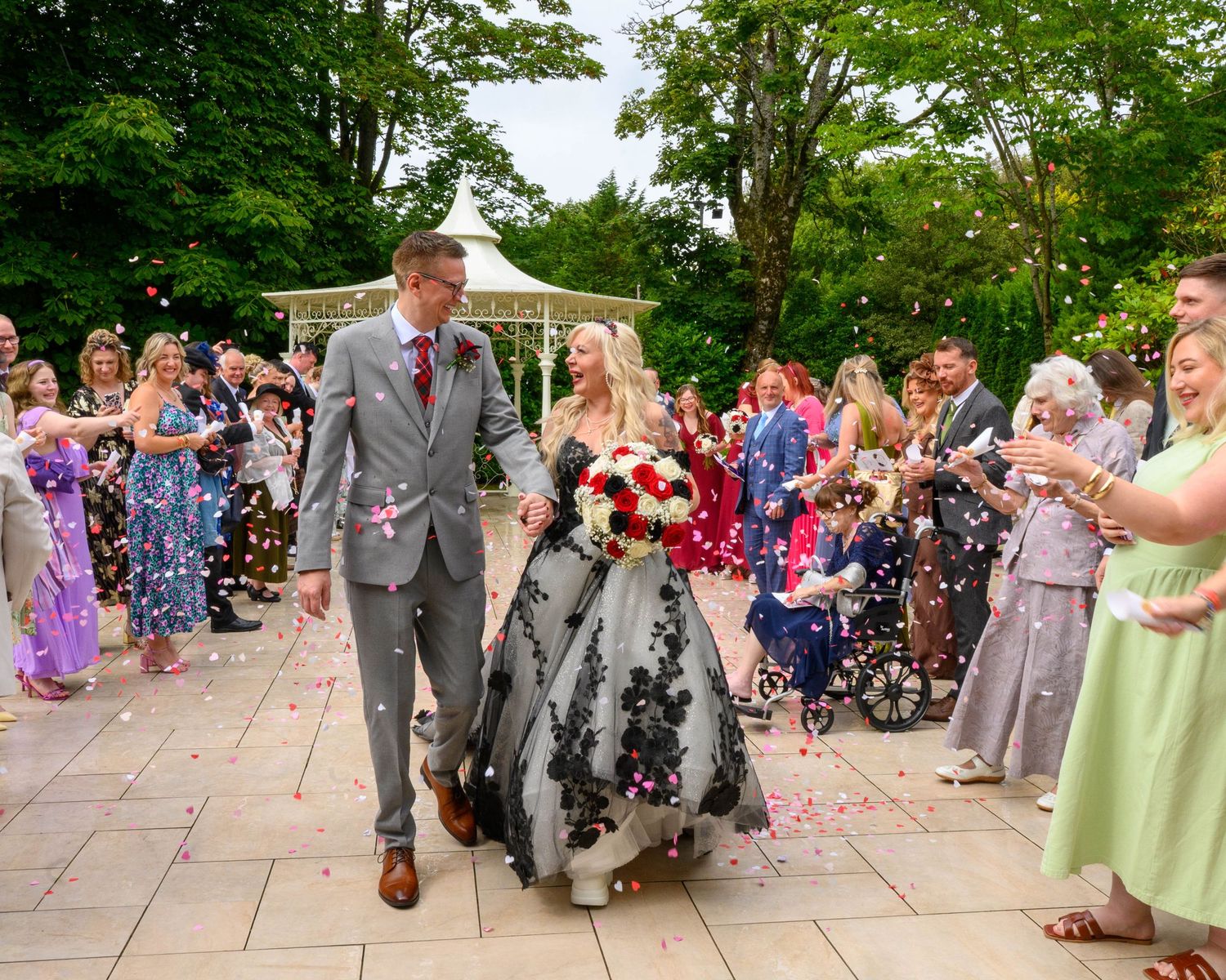 Bride in black lace gown and groom walk through confetti shower at outdoor wedding ceremony.