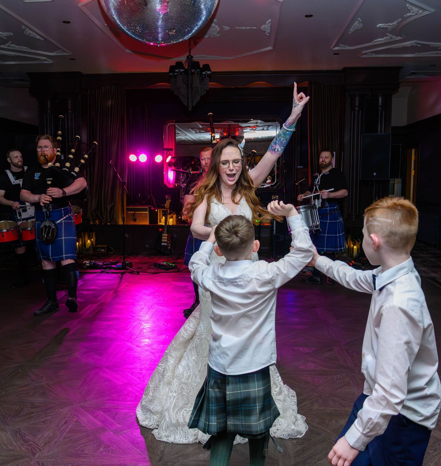 Bride in white gown dances with children in kilts at Scottish wedding reception with live band performing on stage.
