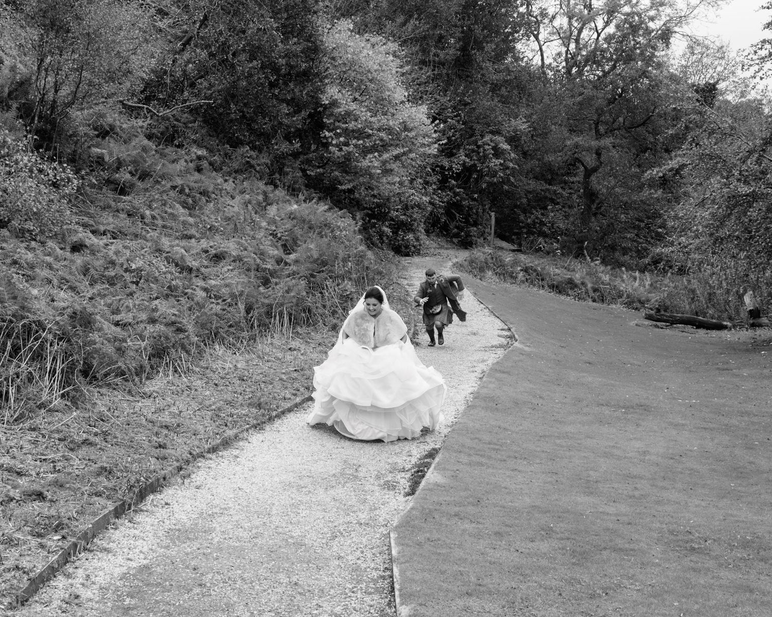 Black and white photo of a bride in a flowing gown walking up a road with guests following behind.