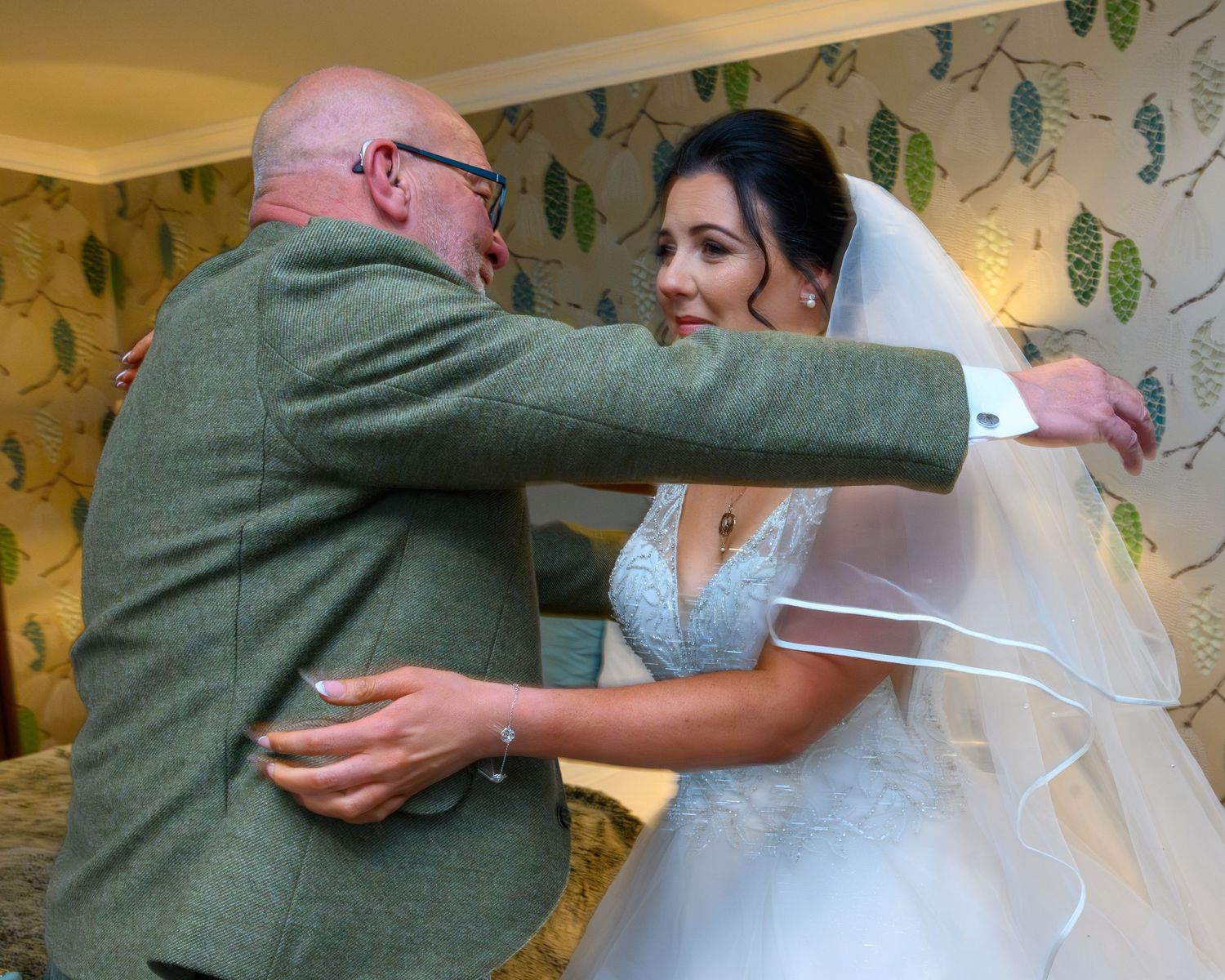 A bride in a white wedding dress embraces someone wearing a green tweed jacket in front of a leafy patterned wallpaper. Gleddoch