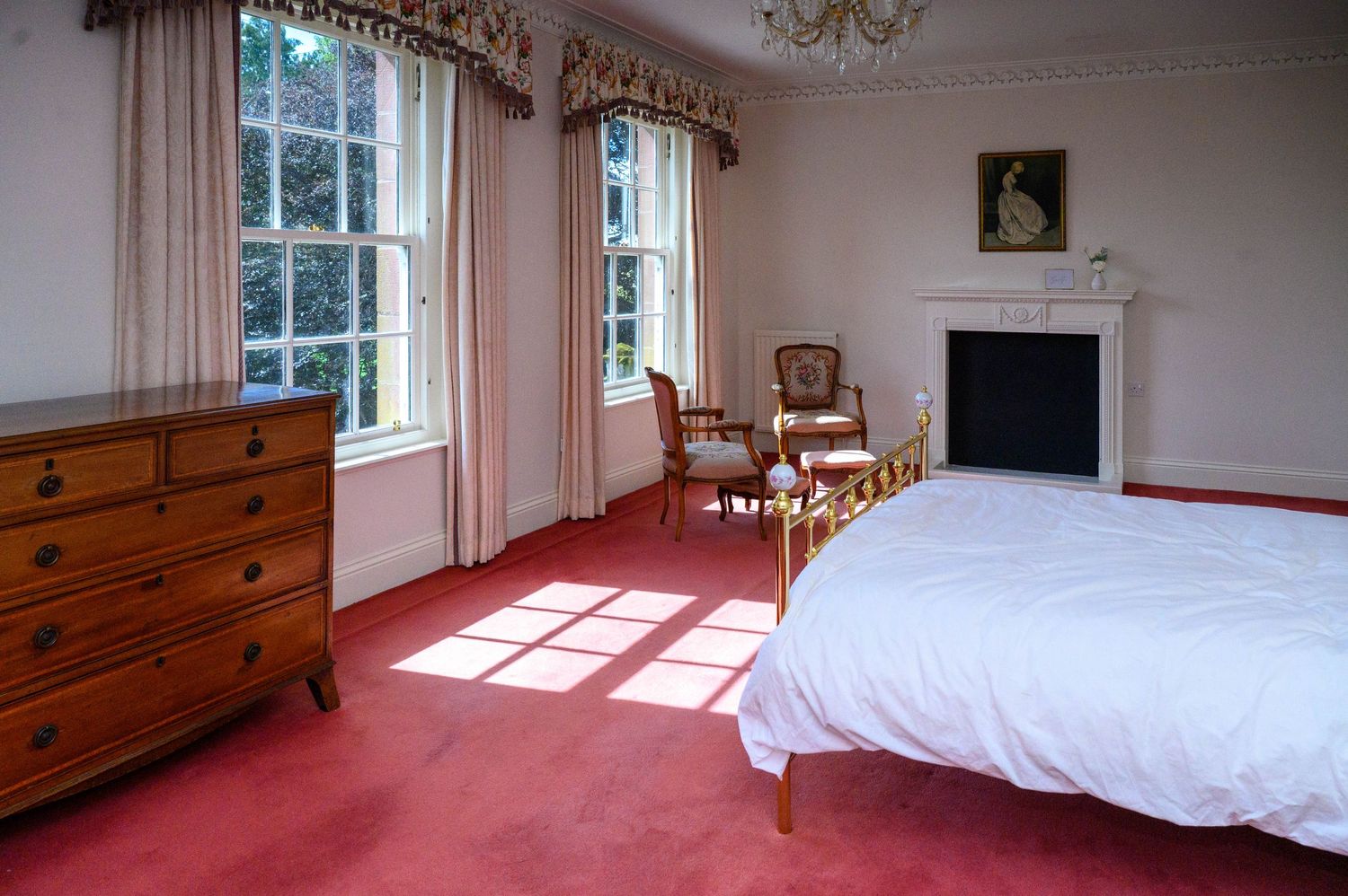 Elegant bedroom with red carpet, white bedding, wooden dresser, fireplace, and tall windows with cream curtains in a period home.