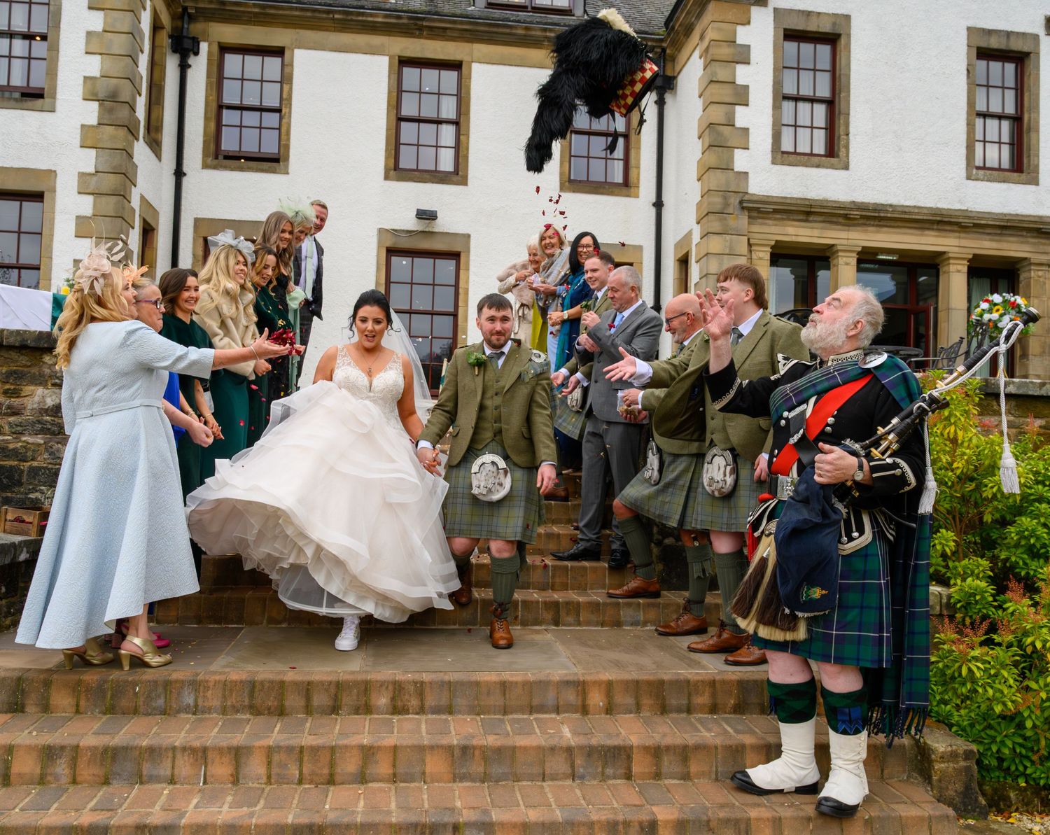 Scottish wedding couple exits venue on steps, greeted by bagpiper in kilt and guests throwing confetti.