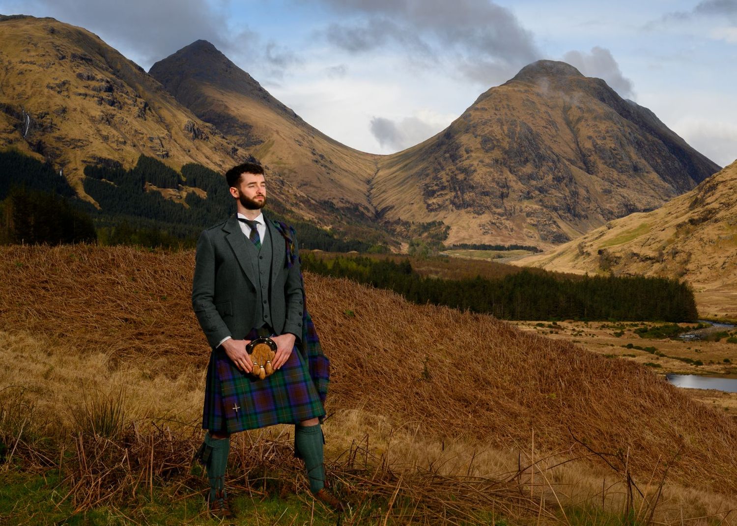 Man in traditional Scottish tartan kilt and tweed jacket standing in Highland glen with mountains backdrop.