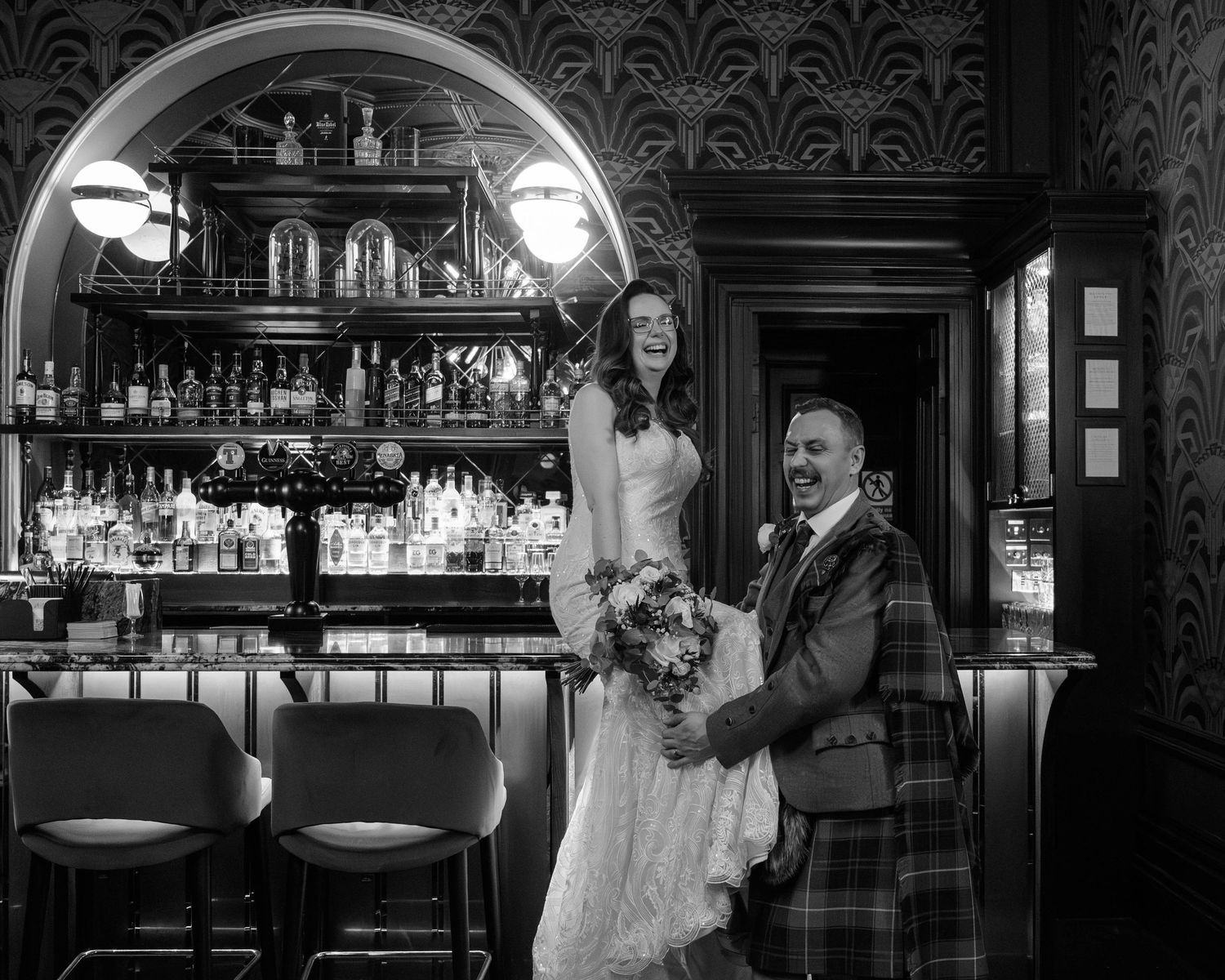 Bride and groom smiling in a stylish bar, groom in kilt holding bouquet, black and white wedding photo.