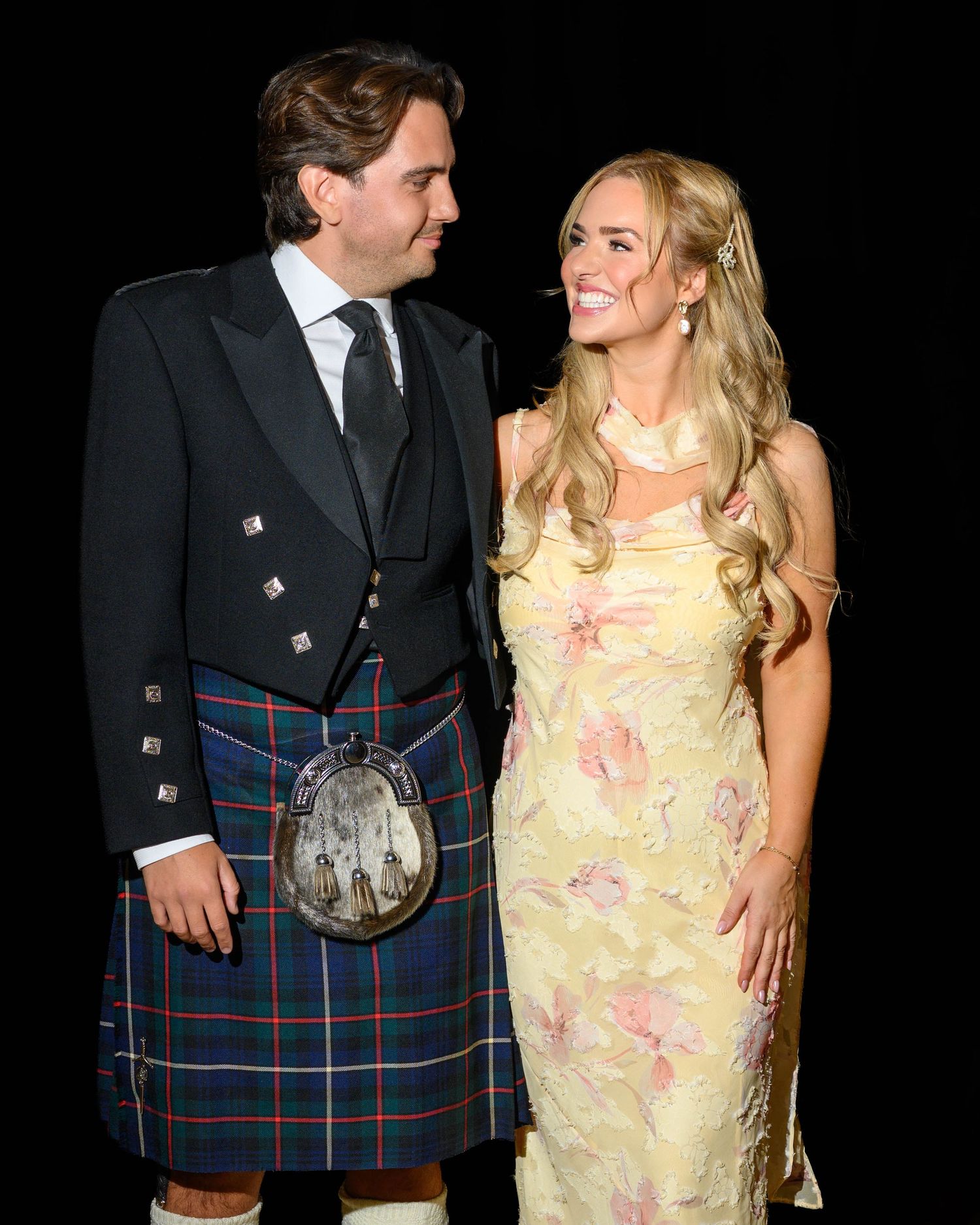 A couple in formal attire, man in Scottish tartan kilt and woman in floral yellow gown, smiling at each other.