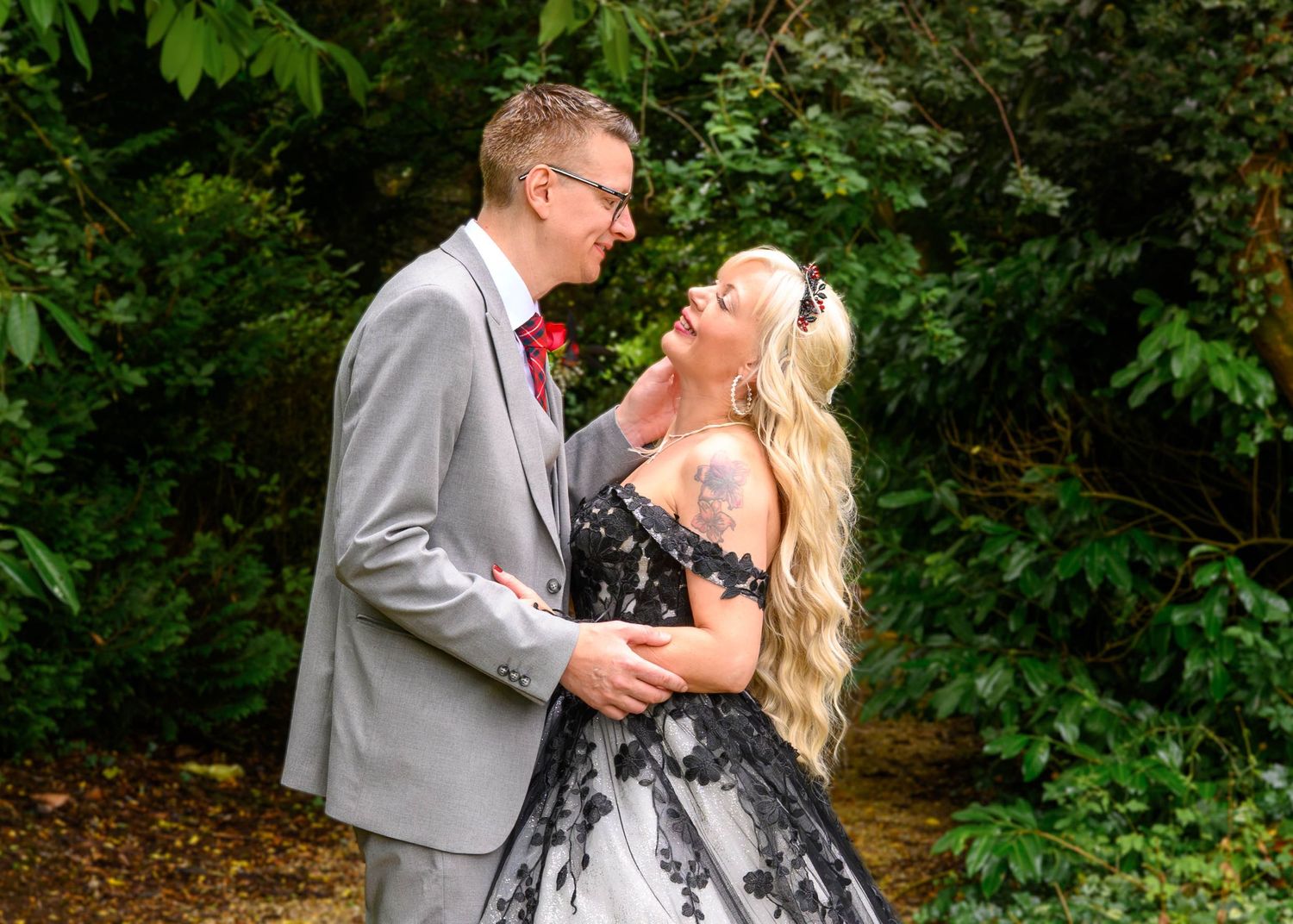 Bride in a black and white lace gown with long blonde hair embraces a groom in a gray suit with a red boutonniere among green foliage.