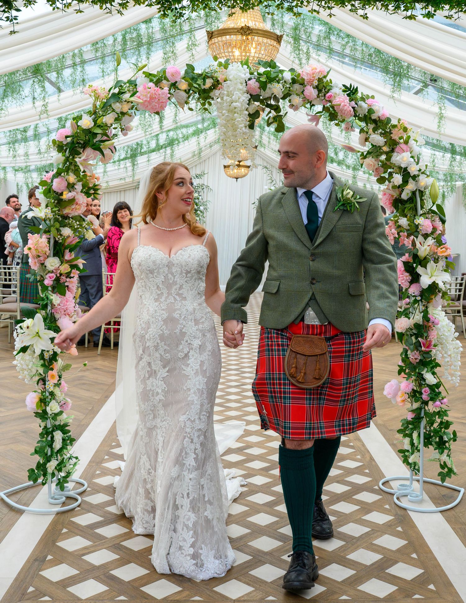 Bride and groom walk hand-in-hand under a floral arch, groom wearing a red tartan kilt at a wedding ceremony.