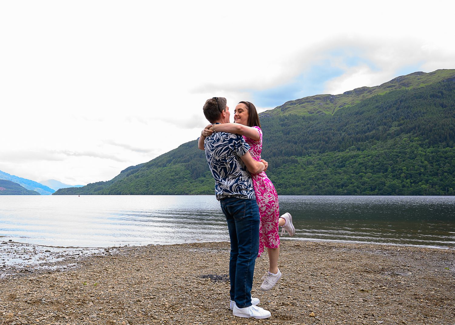 A romantic lift pose by a couple in coordinated outfits against a scenic loch and mountain backdrop. Firkin Point Loch Lomond