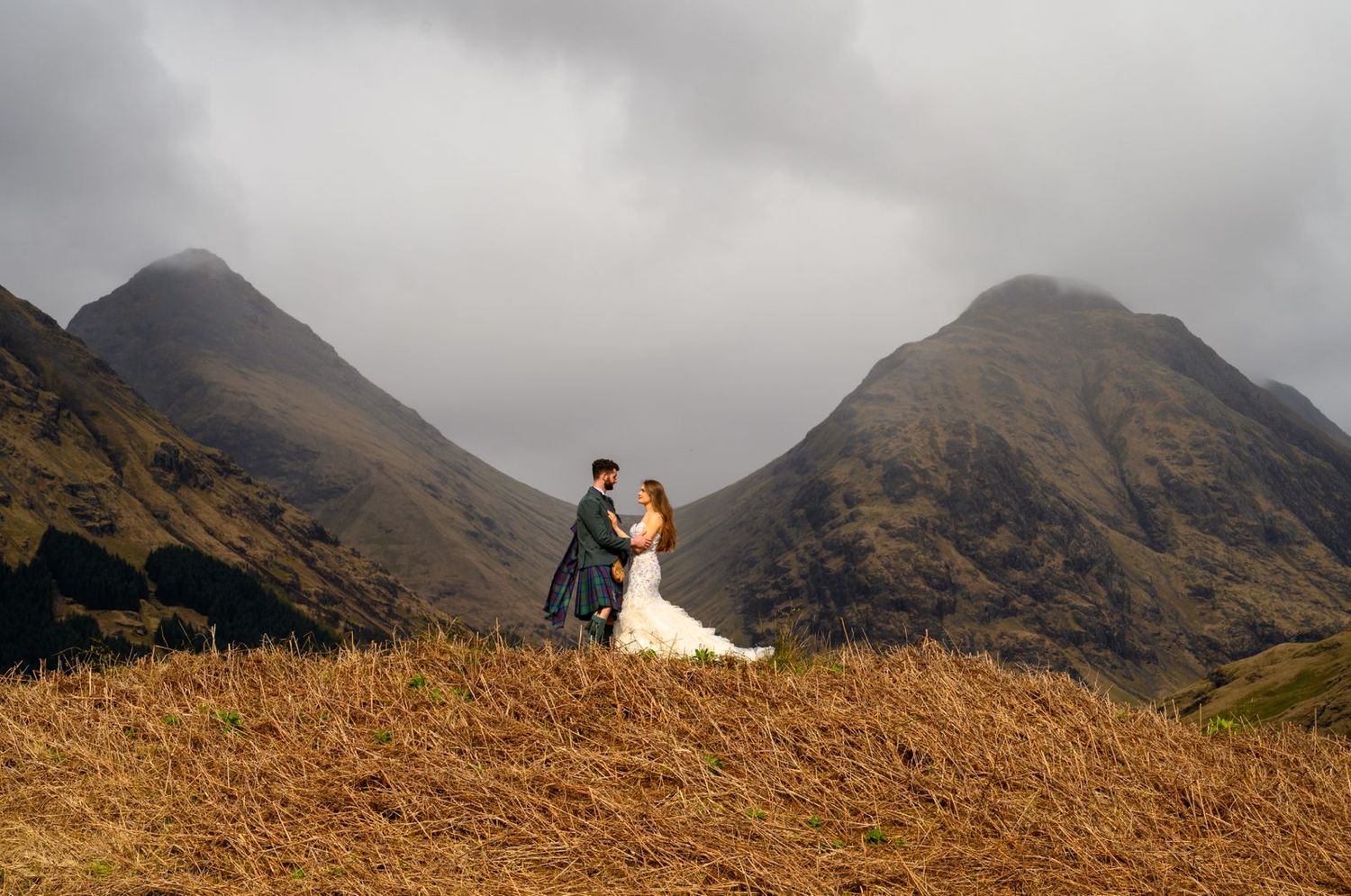 Wedding couple embracing on a golden hillside with dramatic misty mountains in the Scottish Highlands.