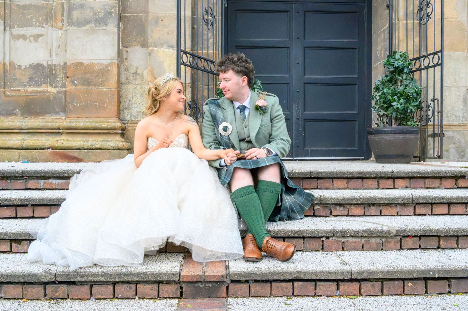 A couple in wedding attire sits on stone steps, the groom wearing traditional Scottish kilt and the bride in a white gown. St Lukes.