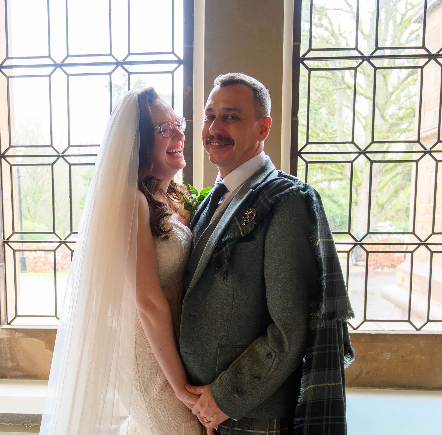 A smiling couple in wedding attire, bride in white gown with veil, groom in tartan kilt, posing before a leaded glass window.