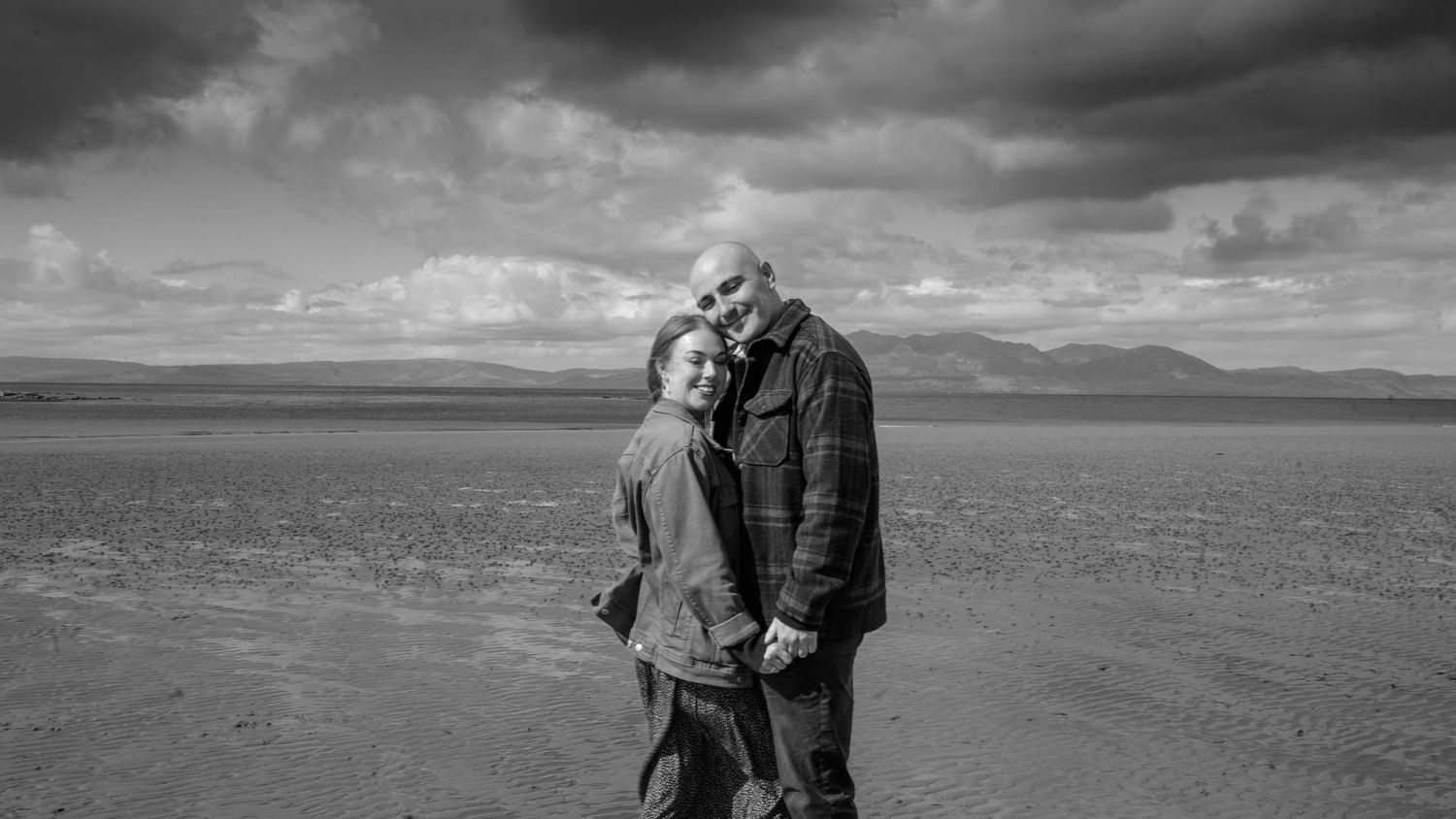 Elderly couple embracing and smiling in a vast open landscape under dramatic cloudy skies, black and white photo.
