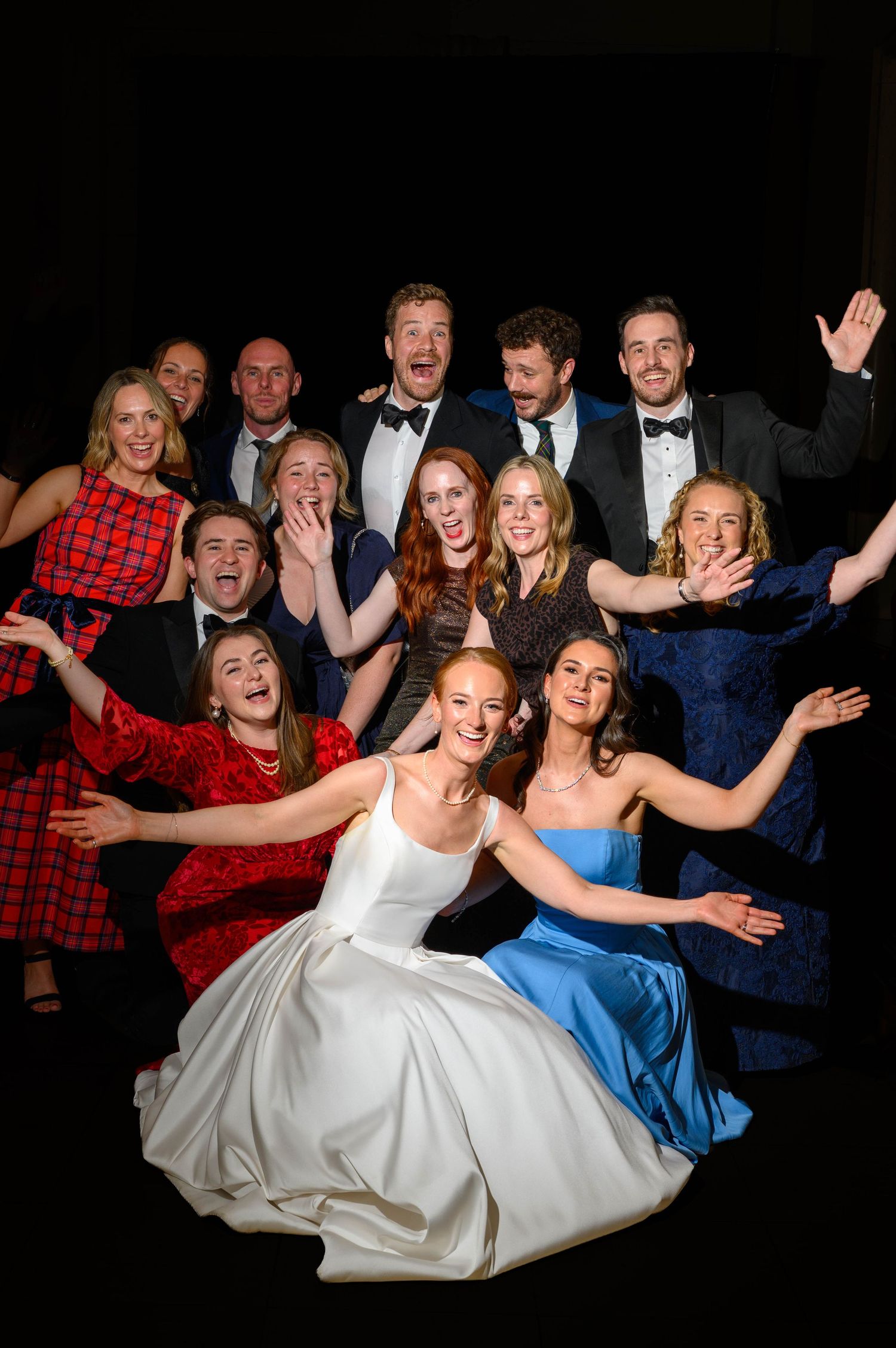 Large group of elegantly dressed wedding guests posing joyfully together, with bride in white gown at center front.