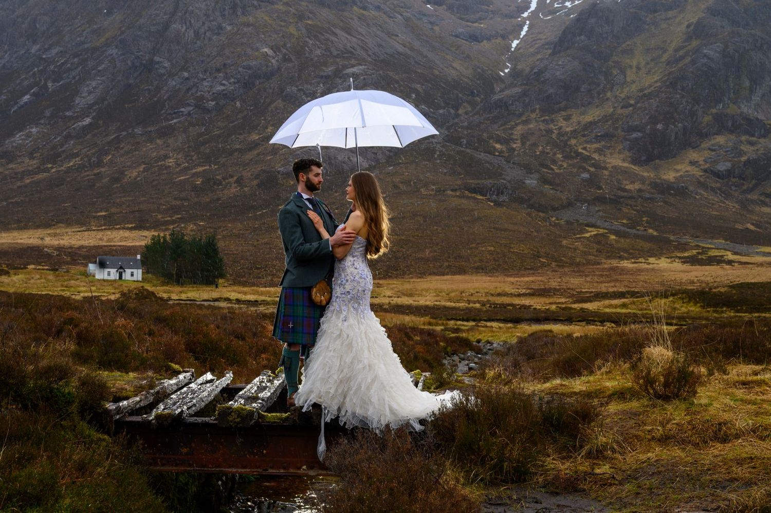 Bride and groom share a romantic moment under a white umbrella in a misty Scottish Highland landscape.