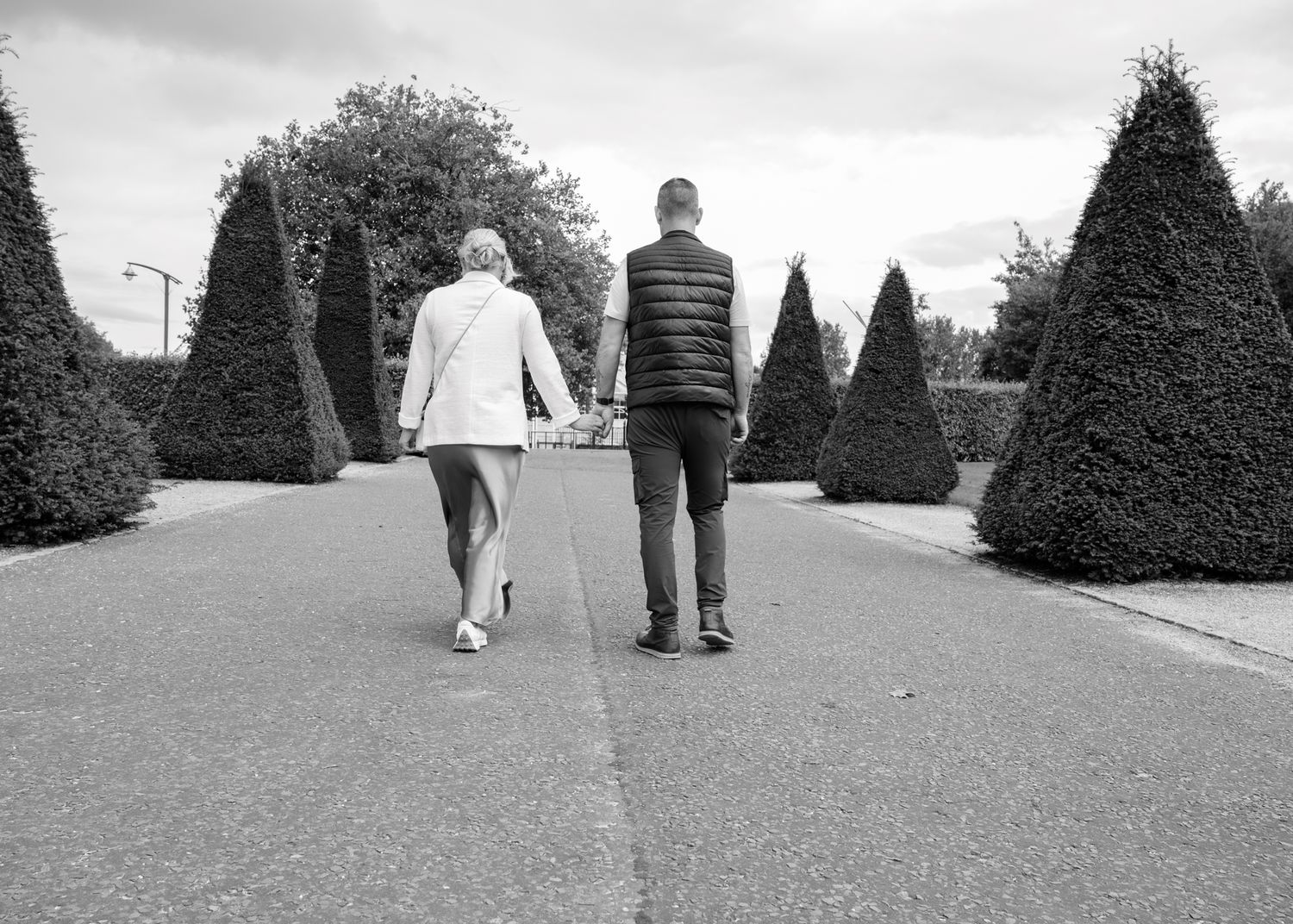 Two people walk along a path lined with perfectly manicured conical topiary trees in a black and white photograph. Glasgow Green