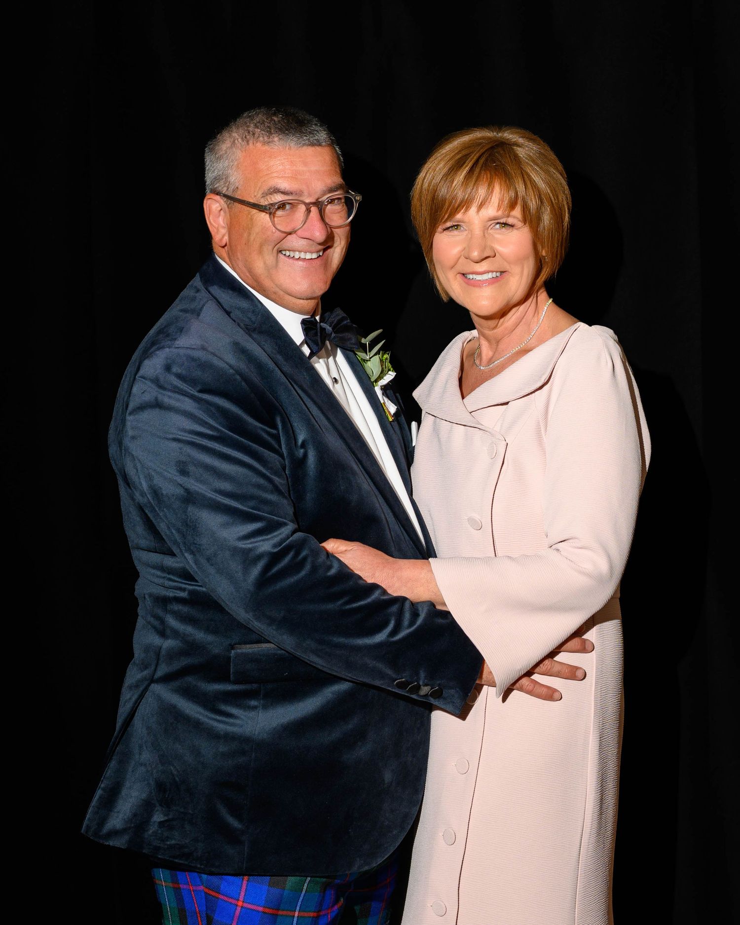 Smiling couple posing together at formal event, man in blue velvet blazer with tartan trousers, woman in cream coat dress.