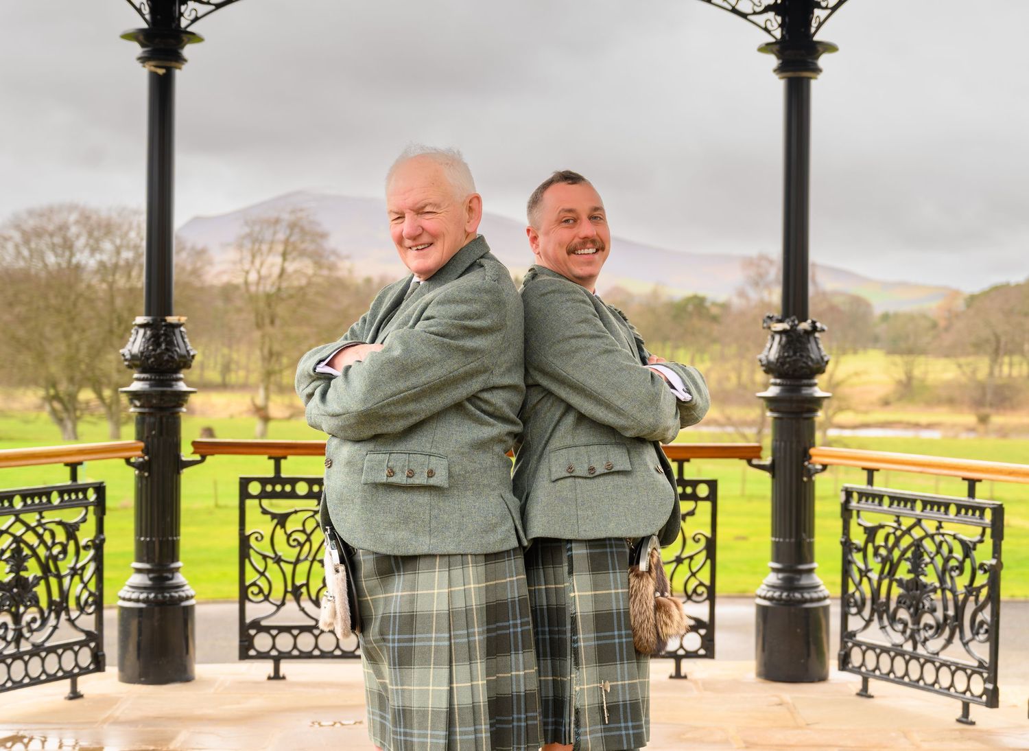 Two men in matching green tartan kilts stand back-to-back smiling under a gazebo with scenic grounds behind them.