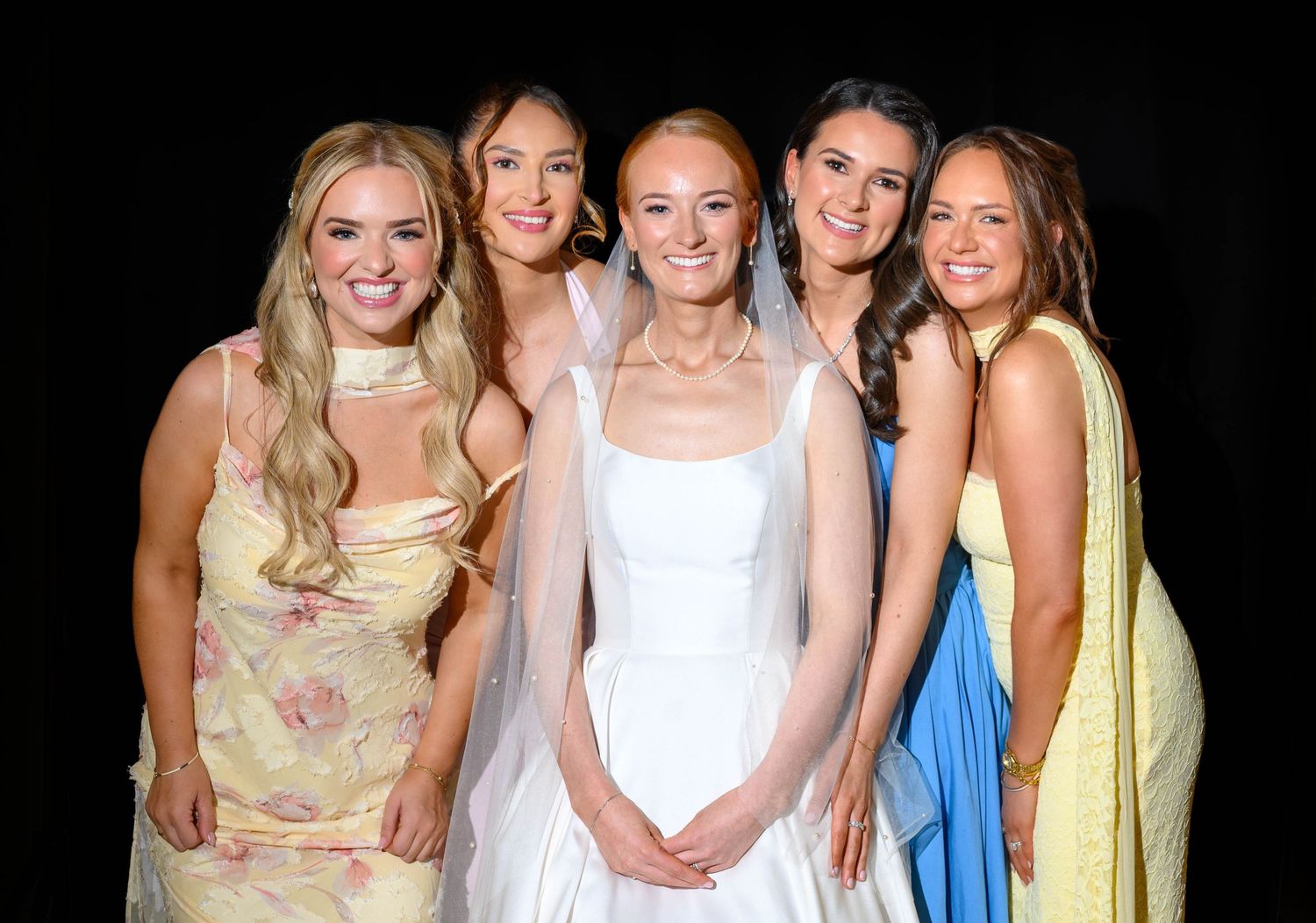 Bride in white gown poses with four bridesmaids in colorful dresses against a dark background.