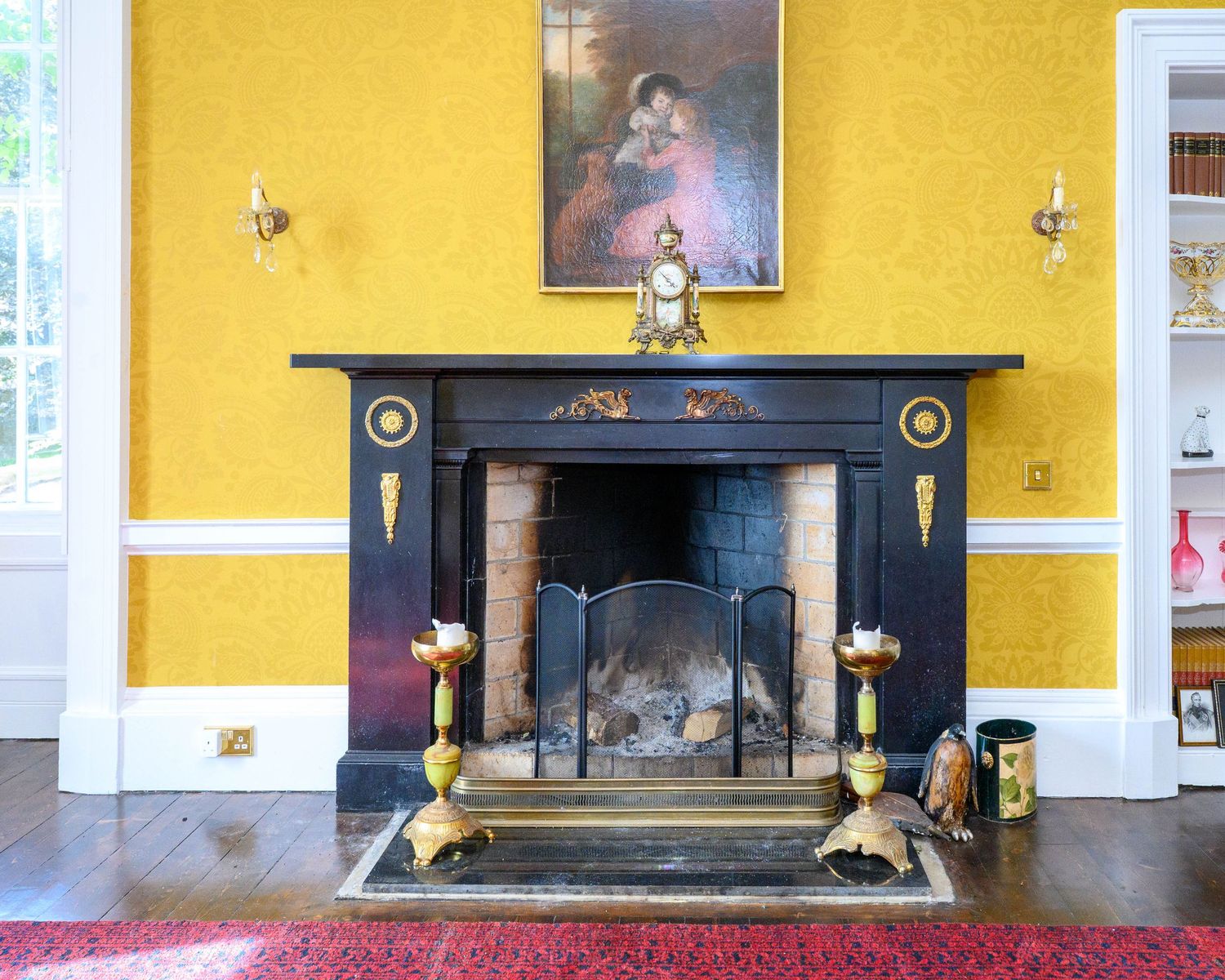 Elegant Victorian fireplace with black surround, gold candlesticks, and painting above on yellow wall.