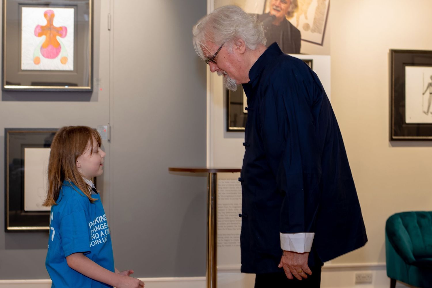 An elderly man in a dark blazer bends down to speak with a young girl in a blue t-shirt inside an art gallery.