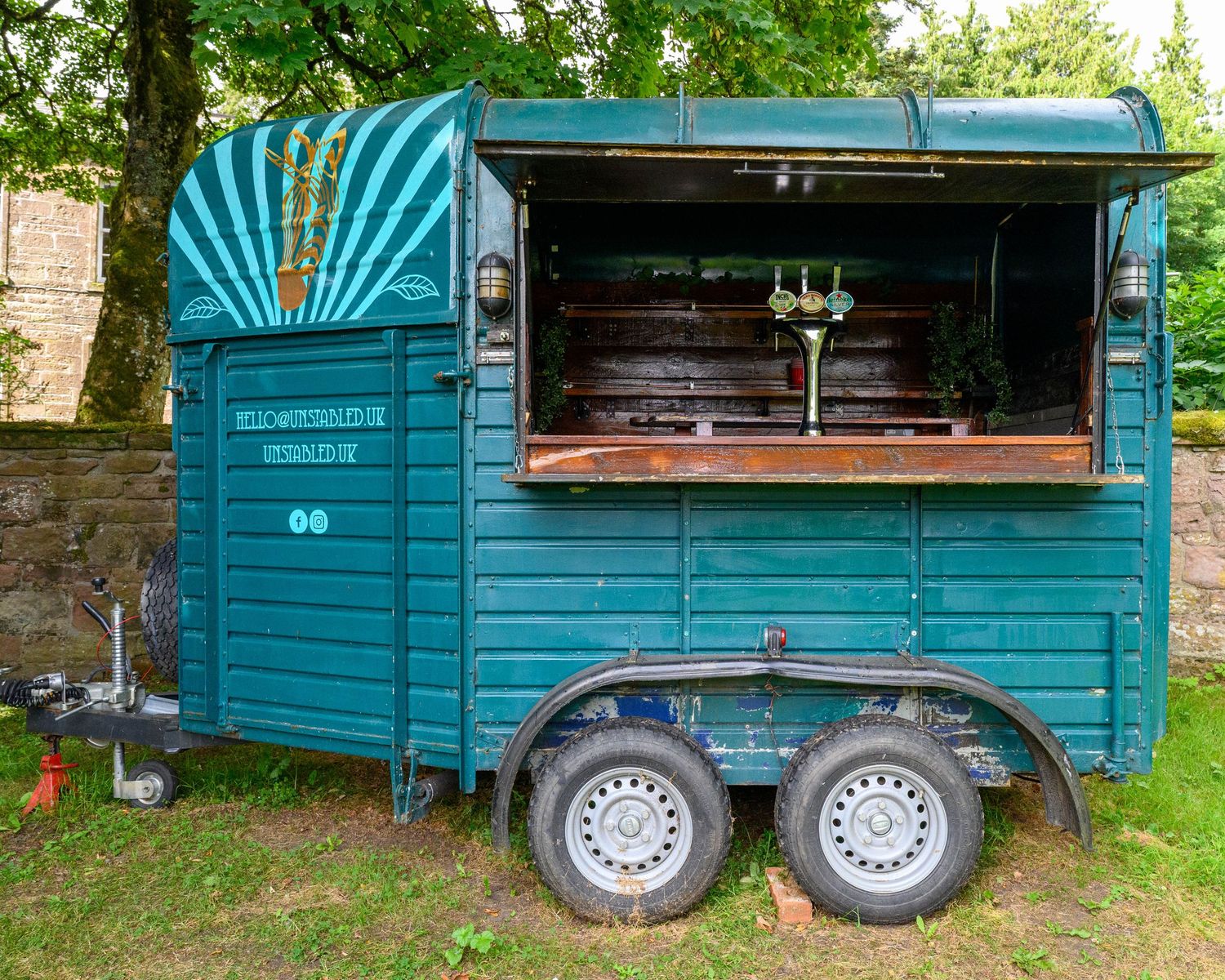 Vintage teal converted horse trailer used as a mobile bar or food stall, parked outdoors near a stone wall.