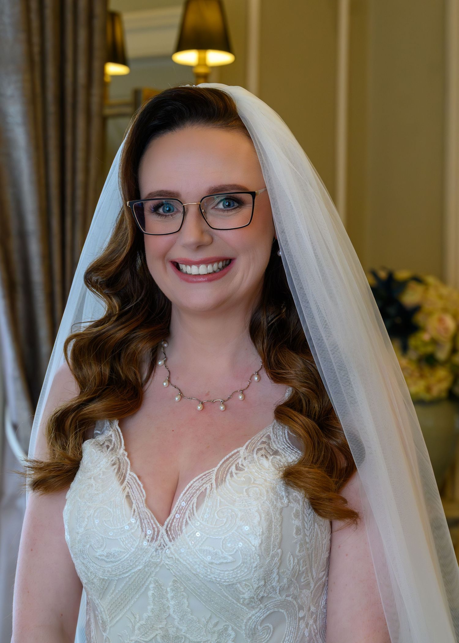 Beautiful bride wearing white lace gown, veil, glasses and pearl necklace smiling indoors.