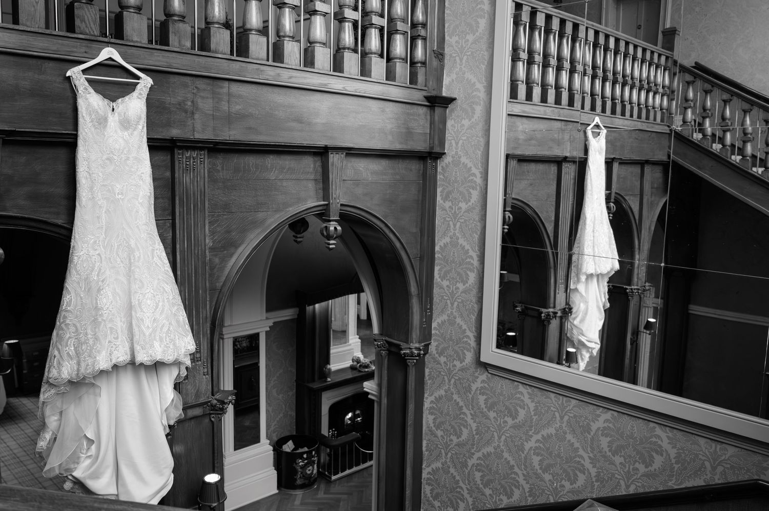 Black and white photo of an elegant wedding dress hanging in a grand wooden staircase hall.