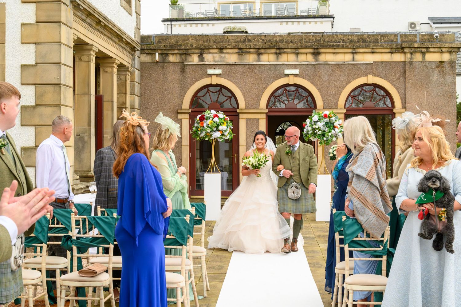 Bride walks down outdoor aisle with groom in kilt at elegant venue, guests applauding, floral arrangements framing the ceremony.