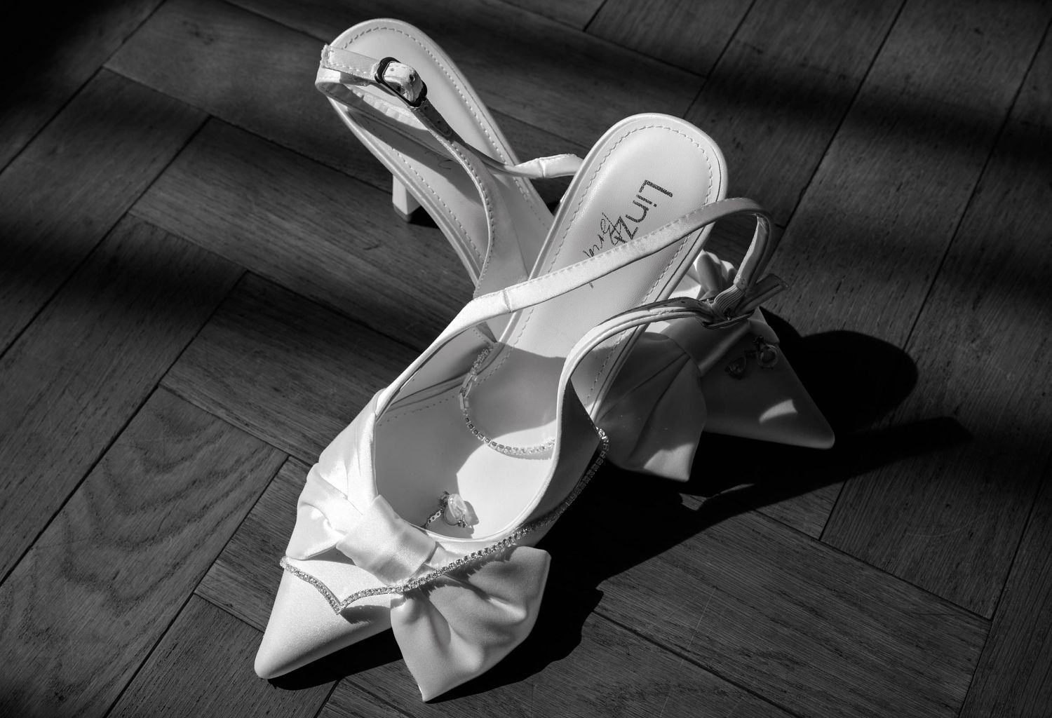 Elegant white satin slingback heels with bow detail on wooden floor, black and white photography.