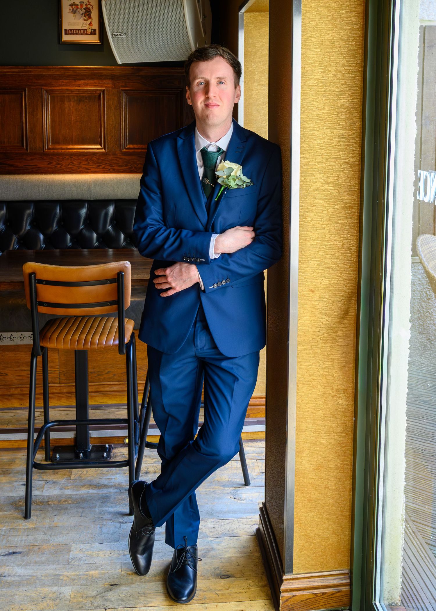 A groom in a navy blue suit with boutonniere leans against a pillar in an elegant venue.