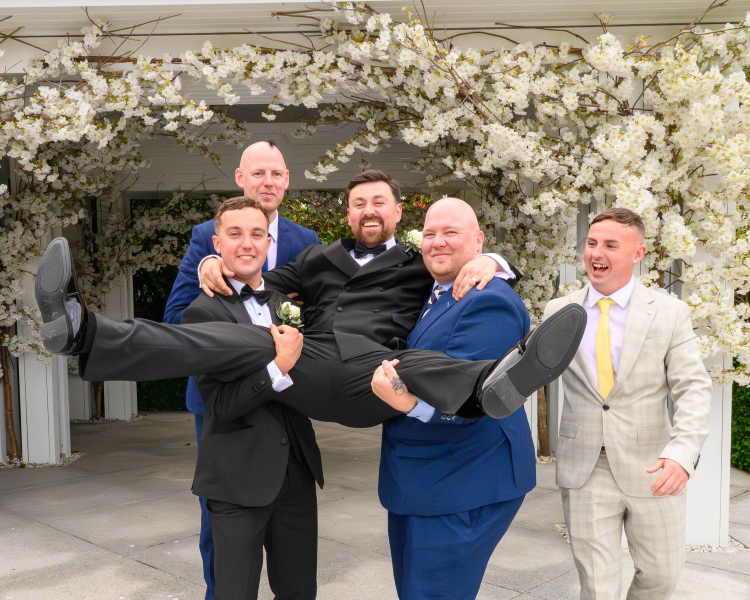 Groom in tuxedo lifted by groomsmen in blue suits under white floral arch at outdoor wedding celebration.