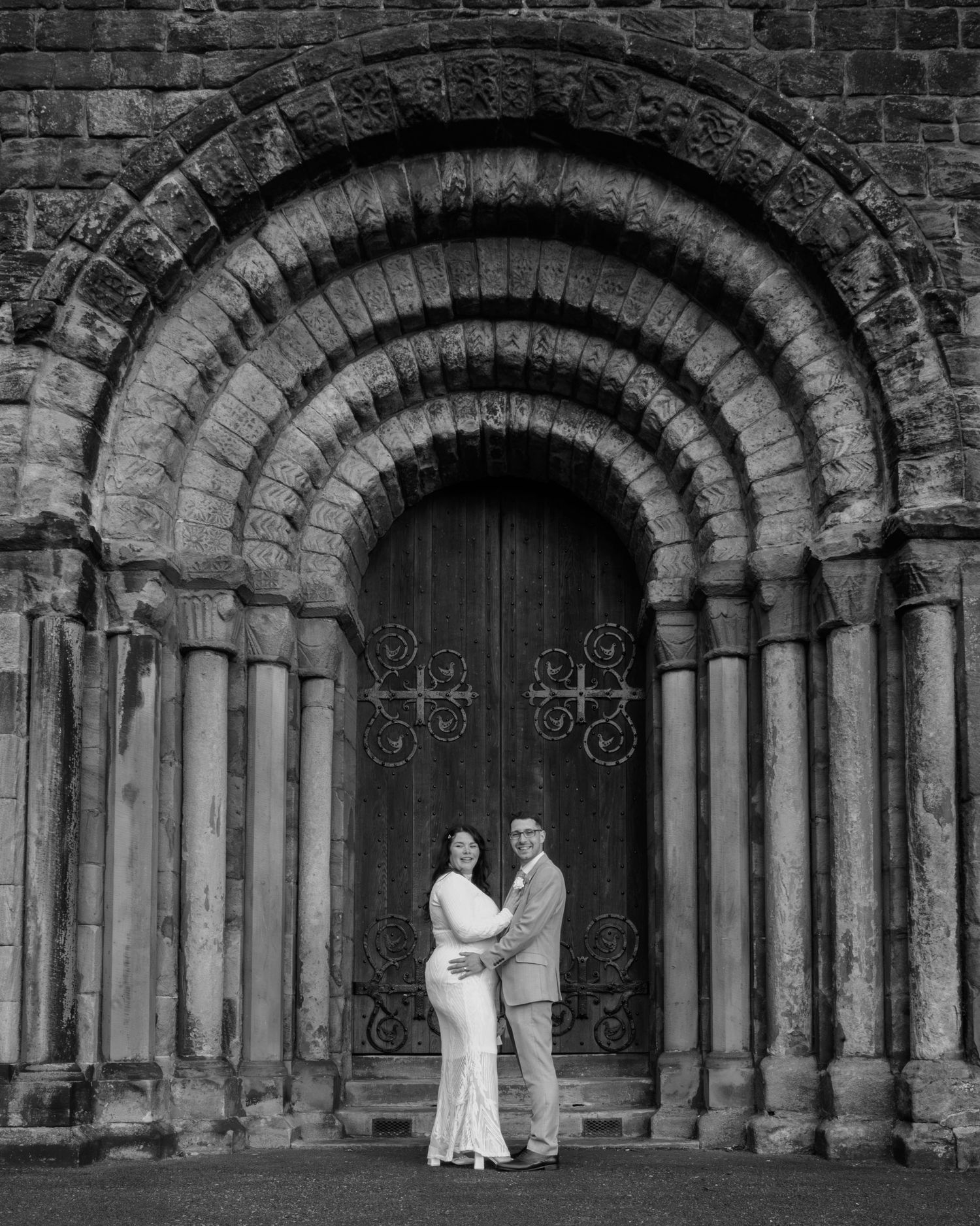 Black and white photo of a couple embracing before an ornate Romanesque stone archway doorway.