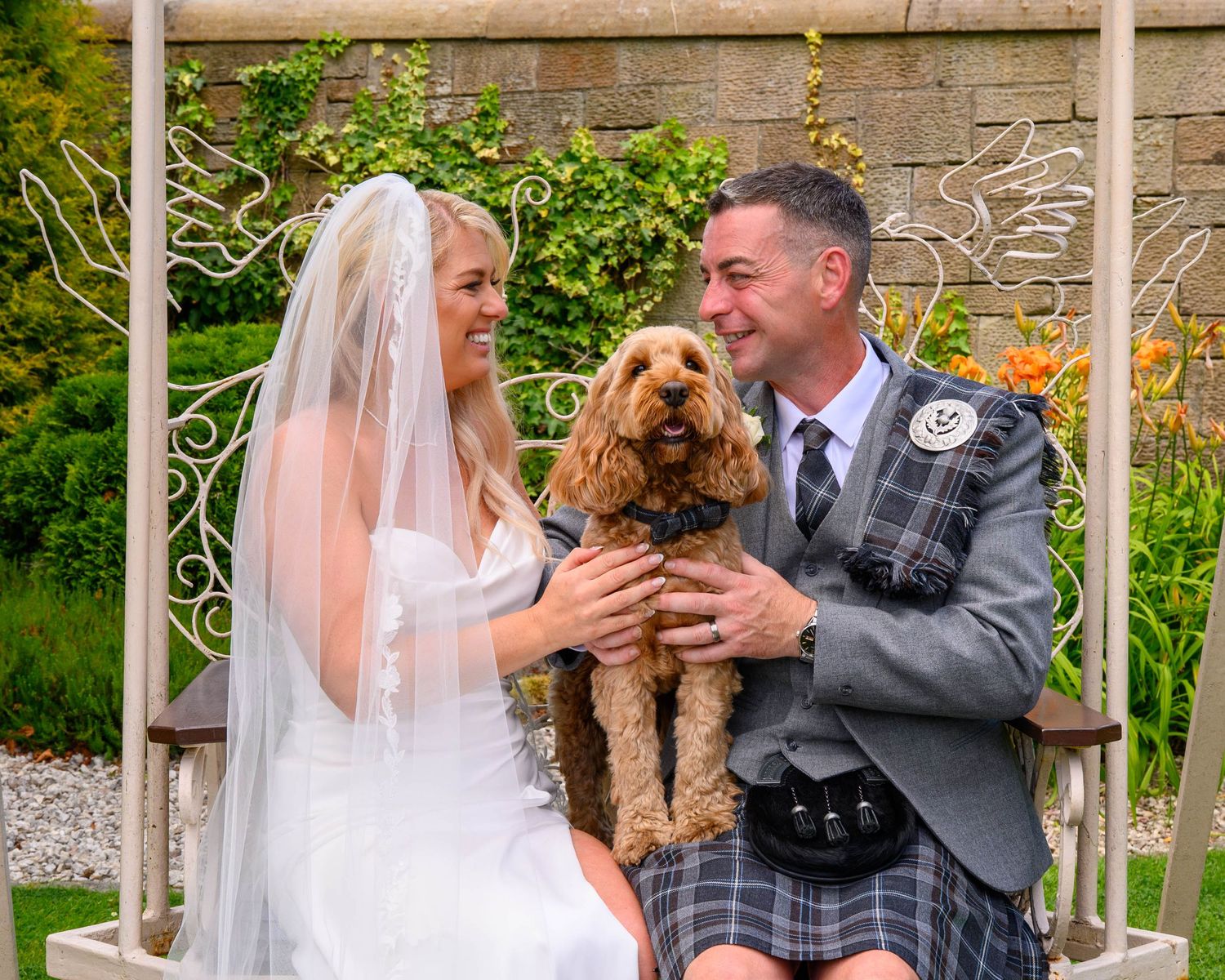 Bride and groom in Scottish kilt sitting on garden swing with cute brown cockapoo dog on their wedding day.