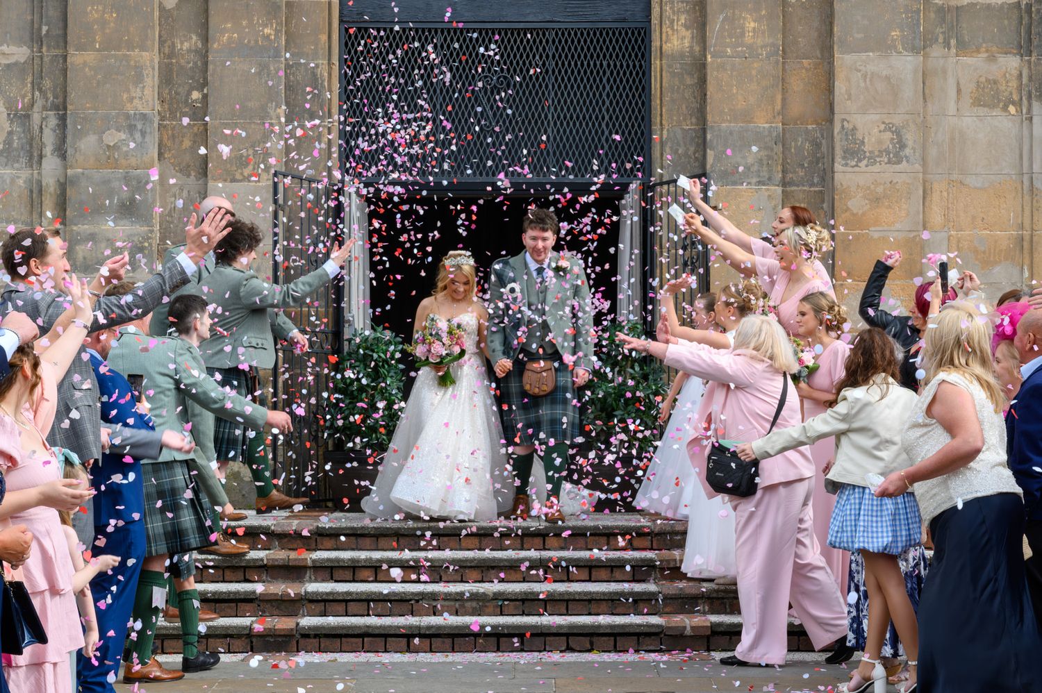 Bride and groom exit church amid colorful confetti shower, surrounded by cheering wedding guests on stone steps.