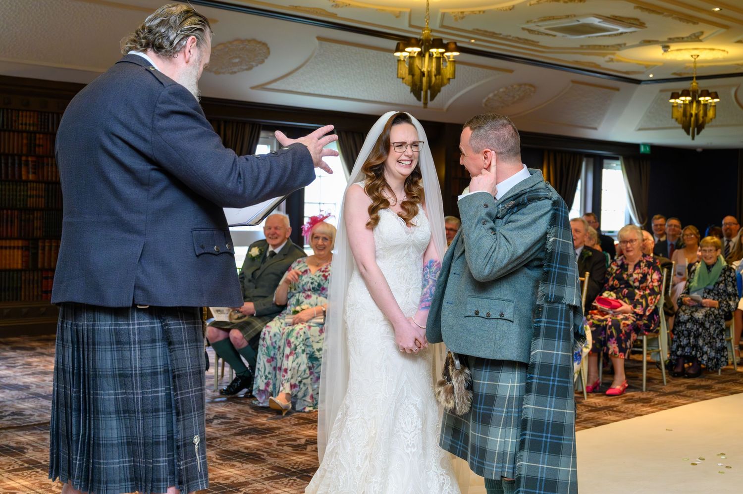 Bride and groom exchange vows in Scottish kilts during elegant indoor wedding ceremony with guests seated behind.