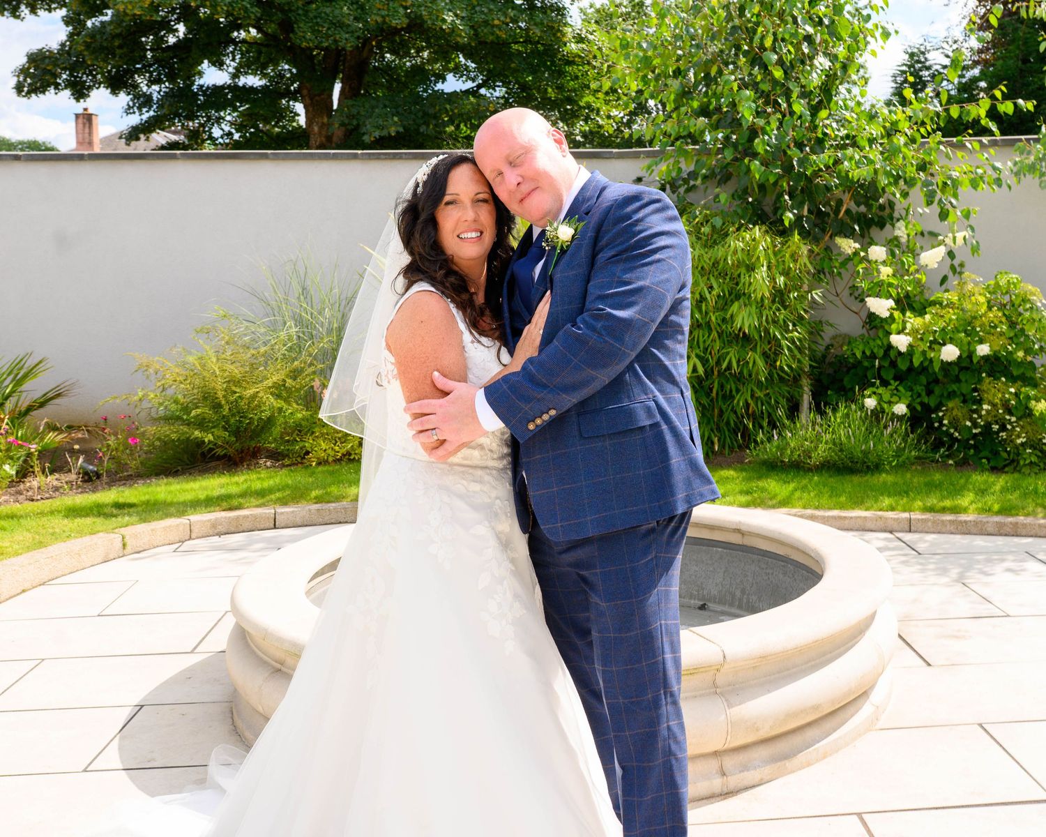 A bride and groom pose together in a sunny garden, she in a white gown with veil, he in a navy blue suit.