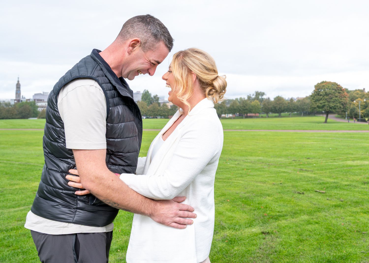 Couple embracing and smiling at each other on a green park lawn with trees in the background.