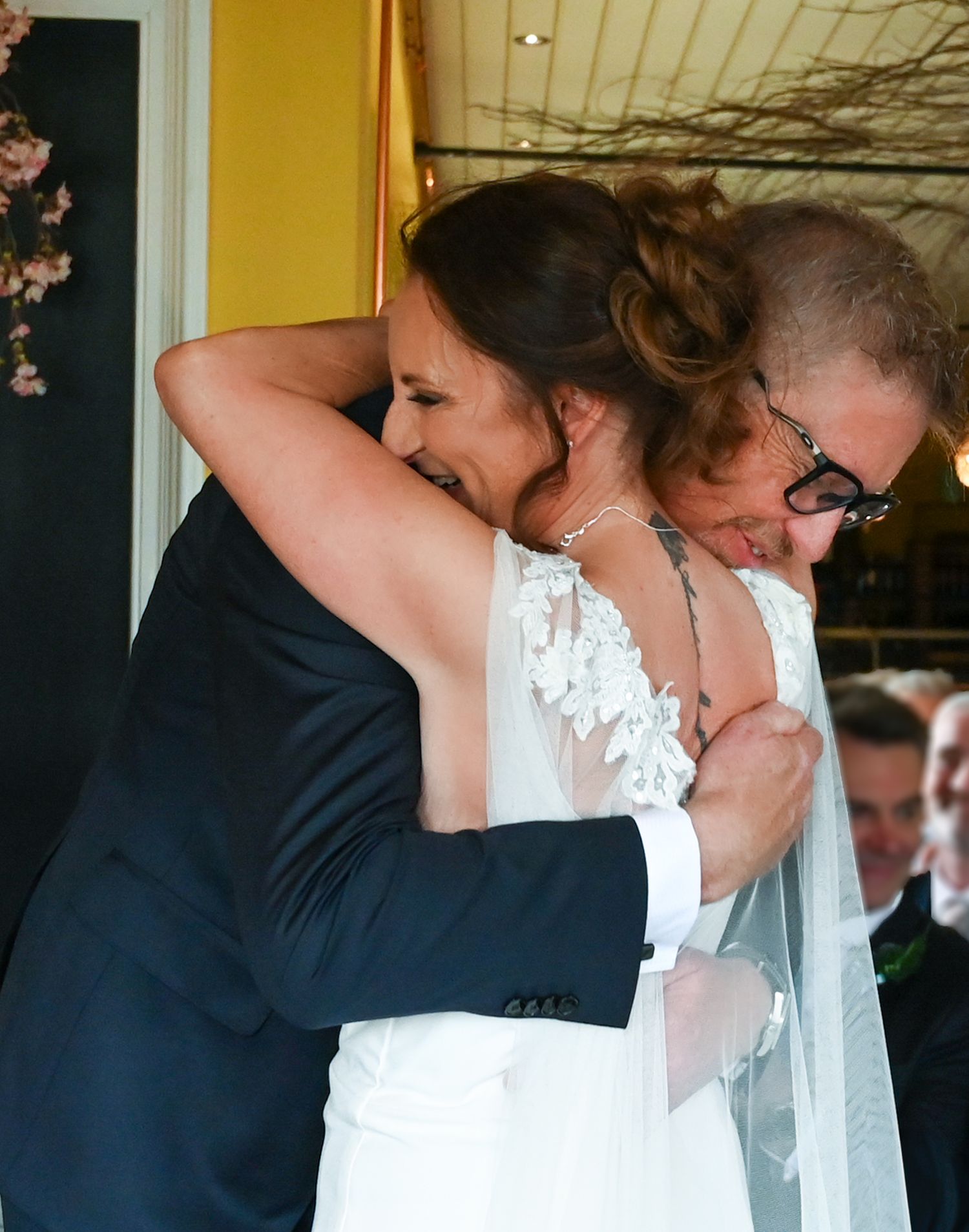 Bride in white lace gown embracing a guest in navy suit at a wedding reception.