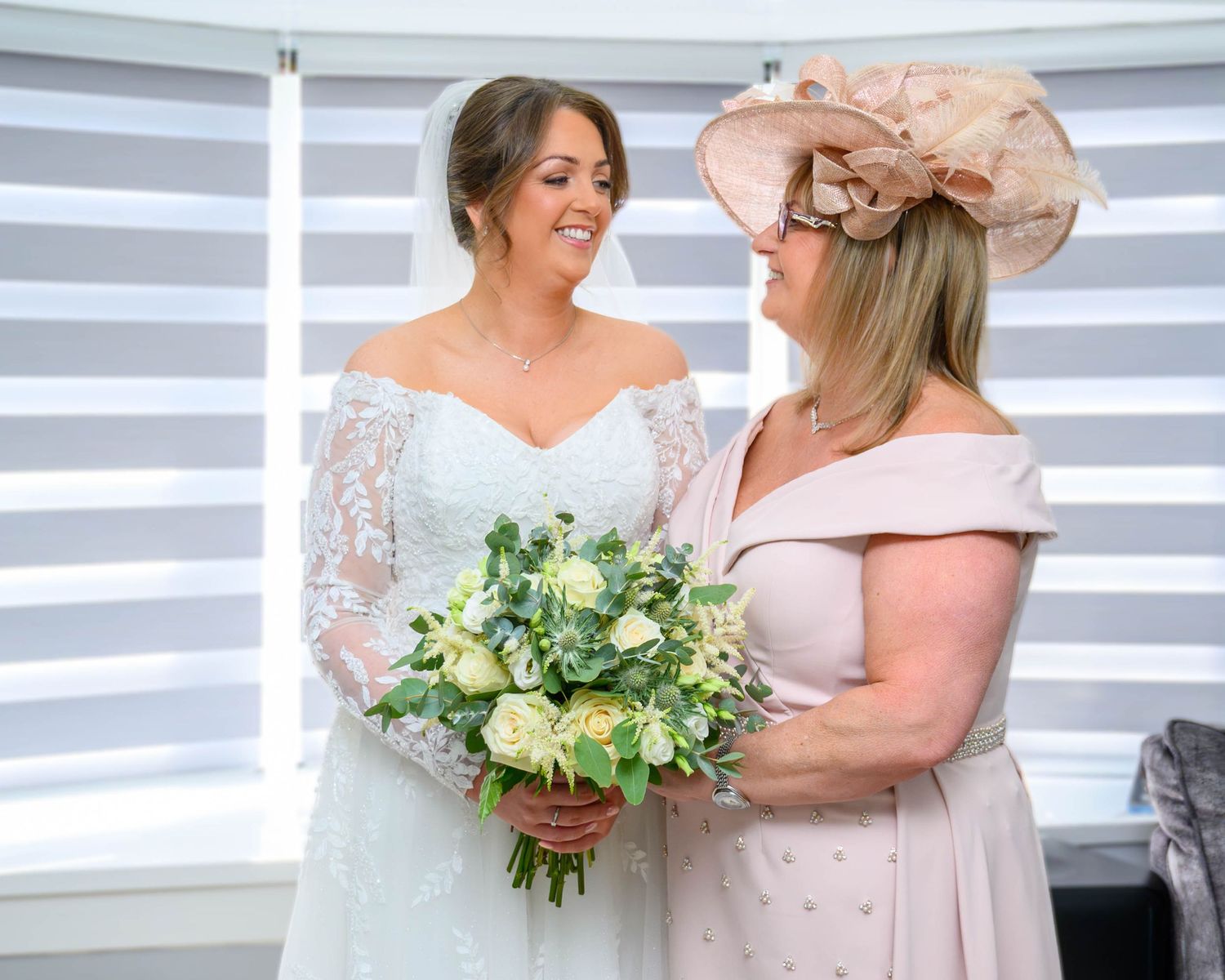 Bride in white lace gown holds bouquet beside woman in pink dress and floral hat near window.