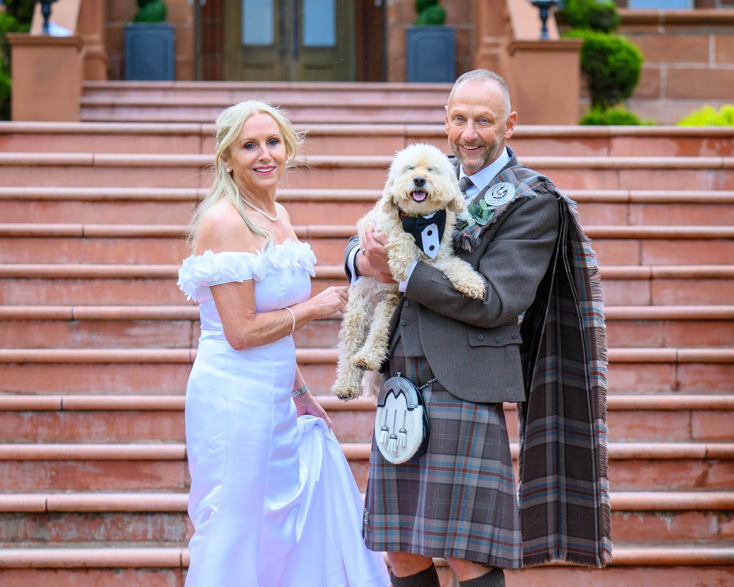 Bride in white gown and groom in Scottish tartan kilt holding a golden doodle dog on stone steps.